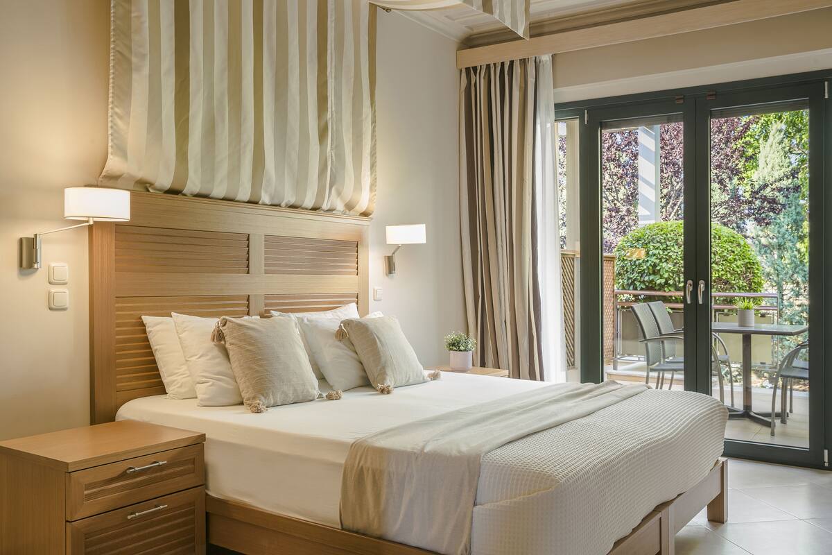 A neatly made bed with a wooden headboard, a bedside table with drawers, and a view of an outdoor balcony with chairs and a potted plant.
