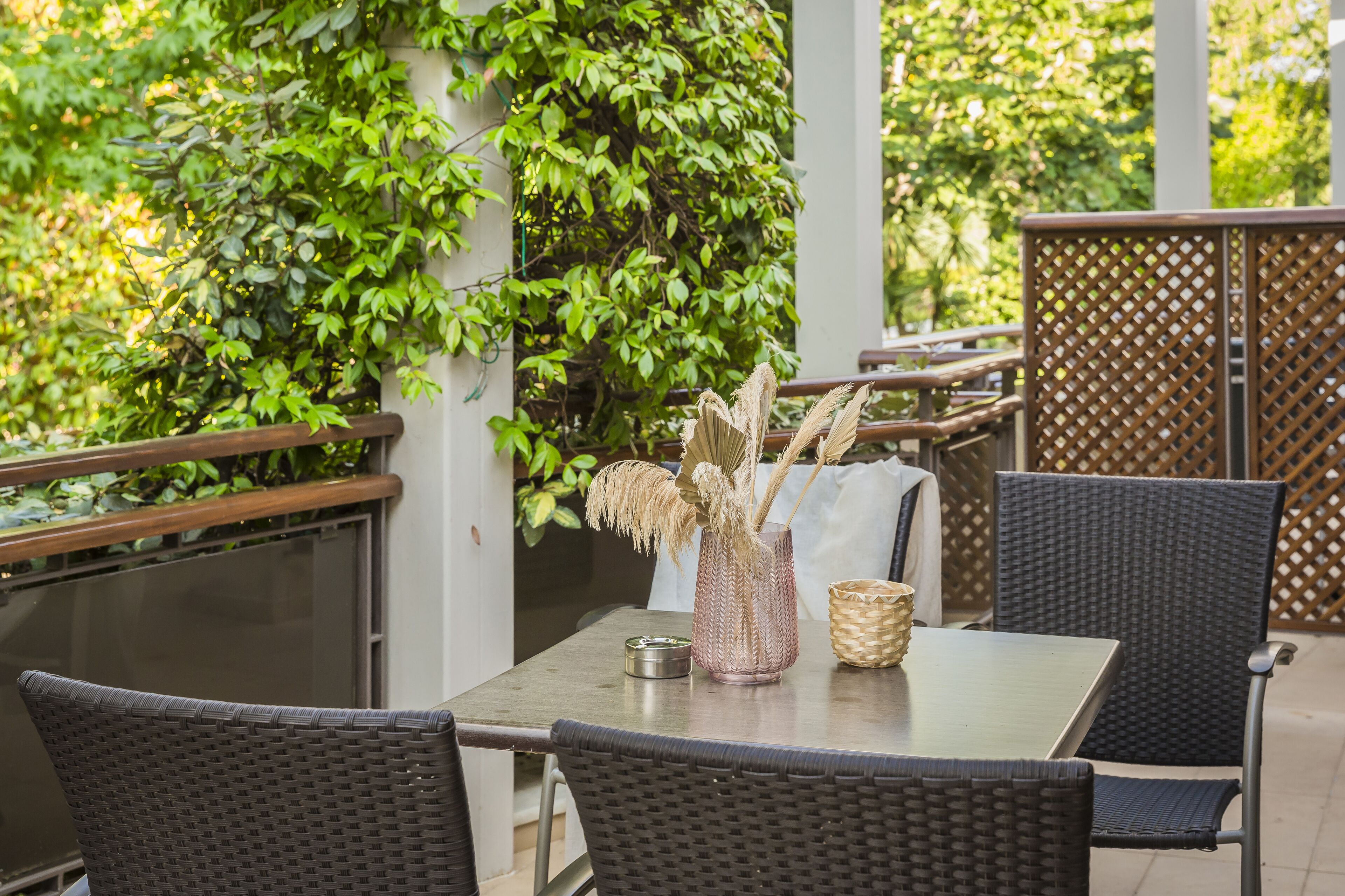 An outdoor patio with wicker furniture, a table with a vase and a metal container, and a backdrop of green foliage.