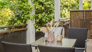 An outdoor patio with wicker furniture, a table with a vase and a metal container, and a backdrop of green foliage.