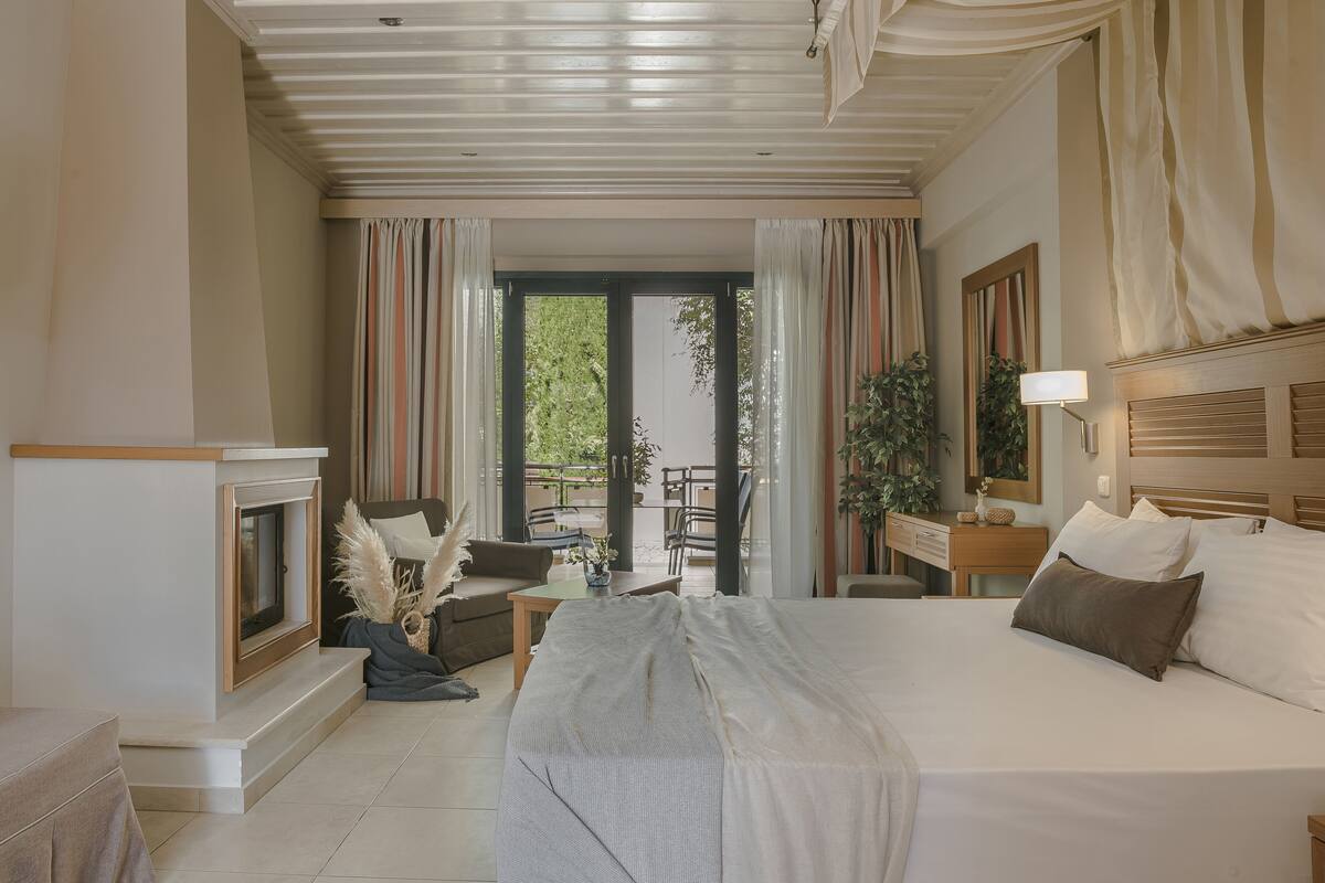 A spacious bedroom with a large bed, a fireplace, a balcony with a table and chairs, and a view of greenery outside.