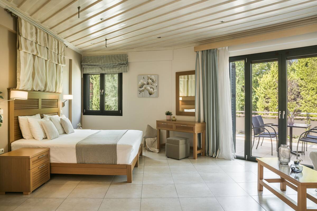 A modern bedroom with a large bed, a wooden desk, a chair, and a balcony with a view of trees.