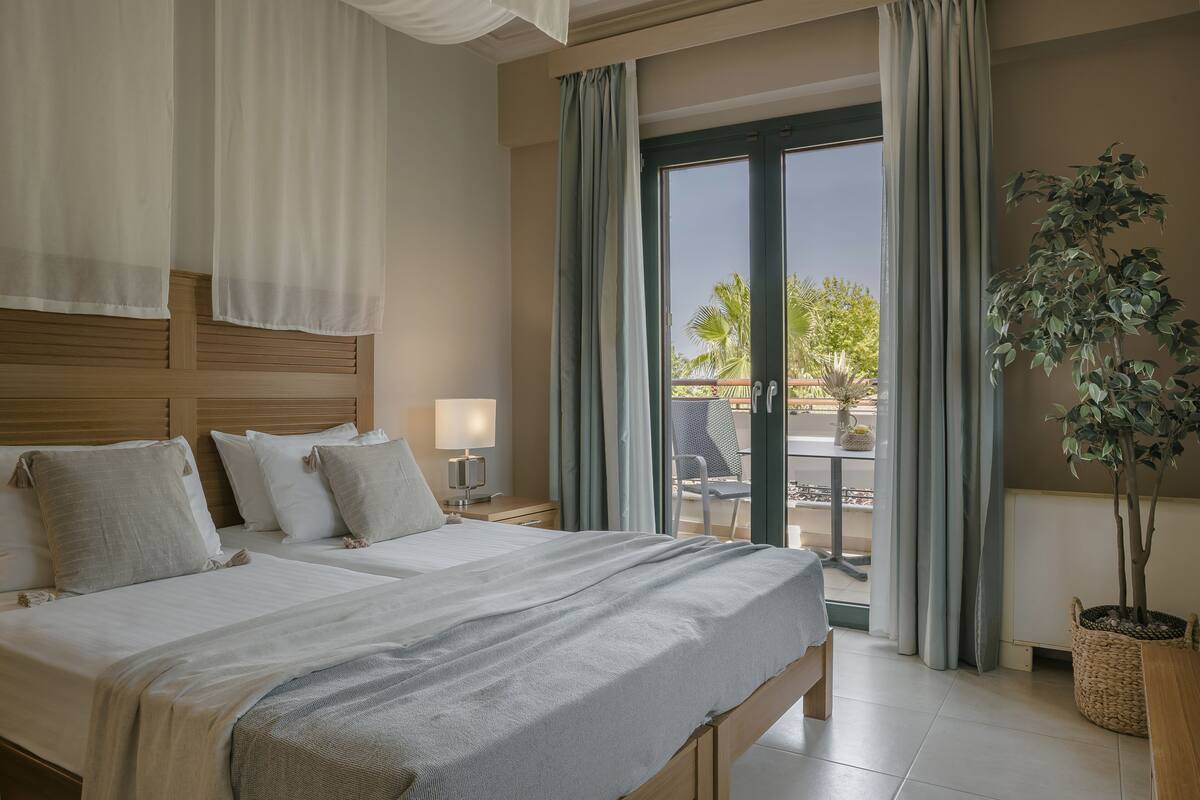 A hotel room with two beds, a balcony with a view, a potted plant, and a table with chairs outside.