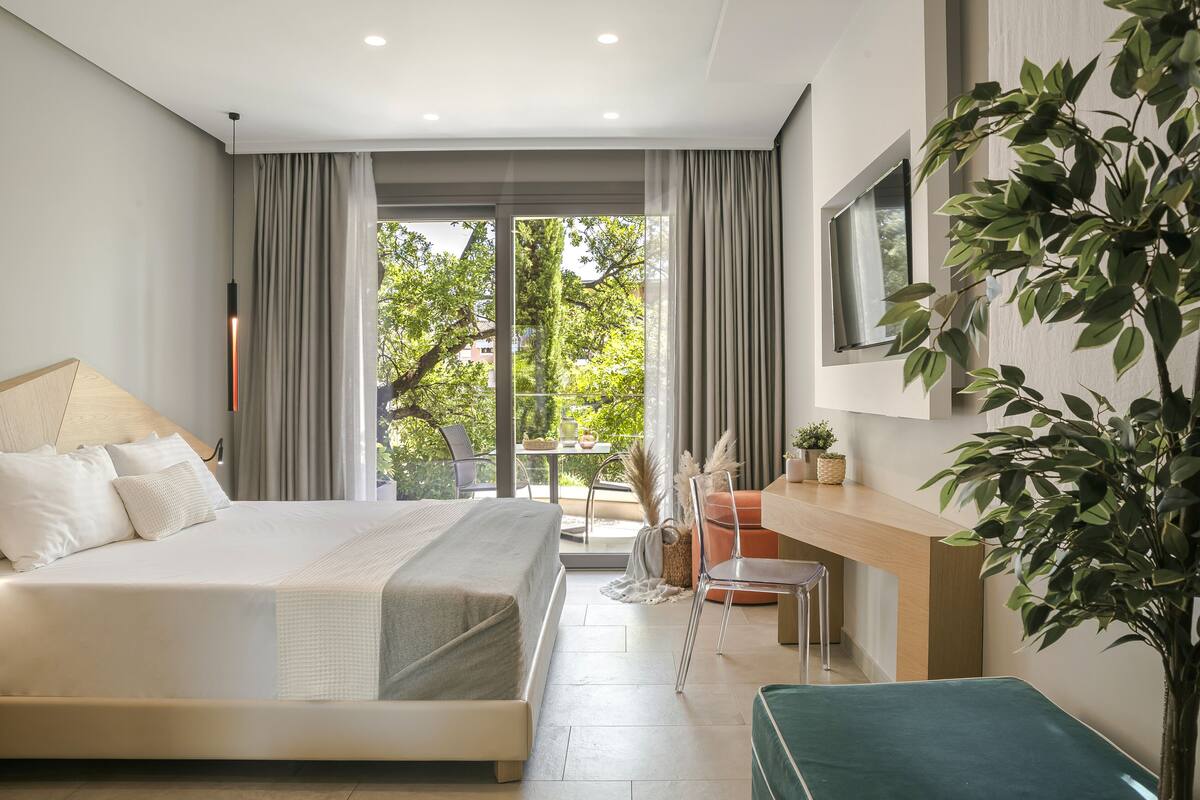 A modern bedroom with a large bed, a desk, and a view of greenery outside.
