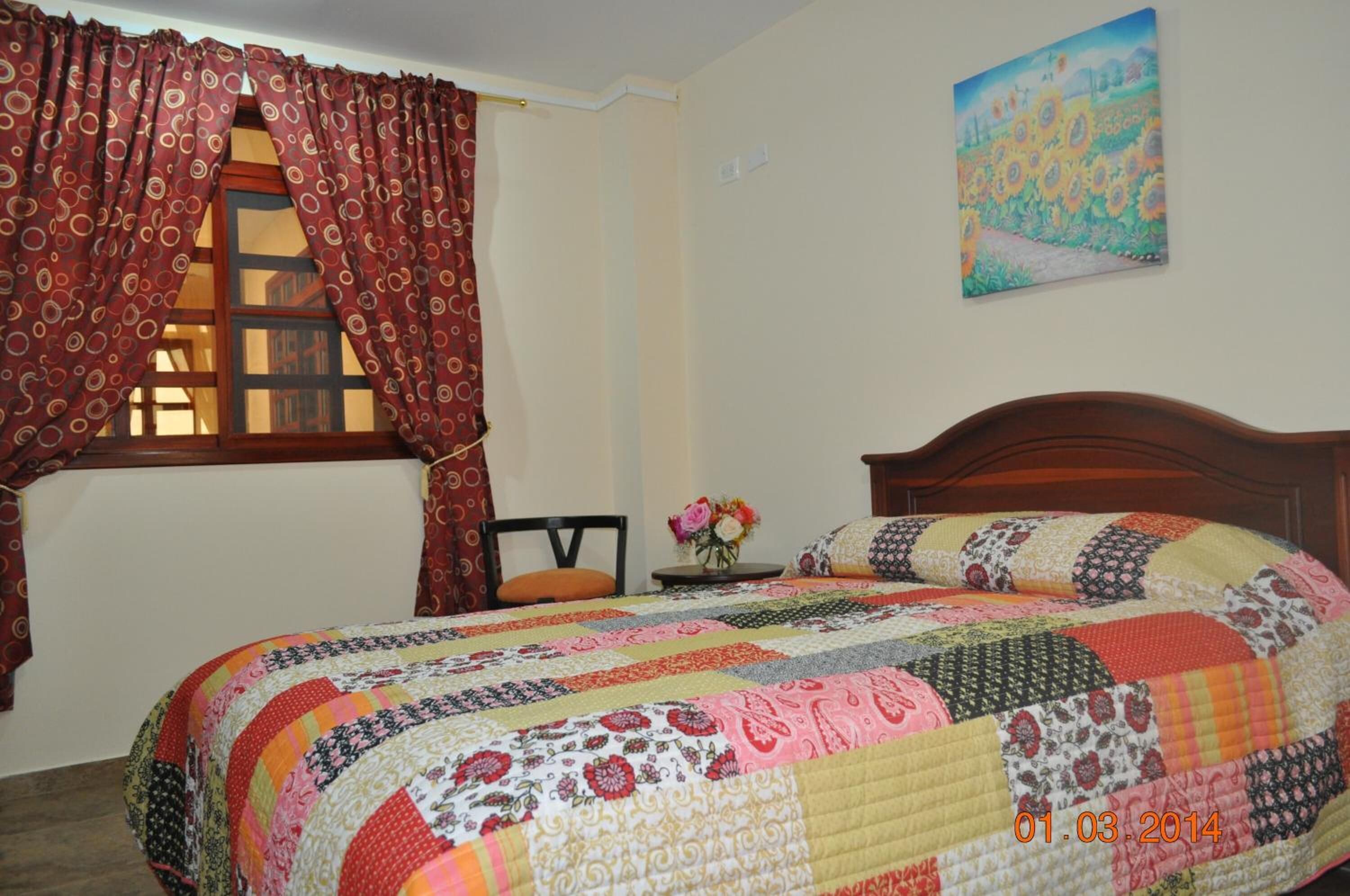Un dormitorio con una cama grande, cabecera de madera, ventana con cortinas, una mesita con un florero y un cuadro en la pared.