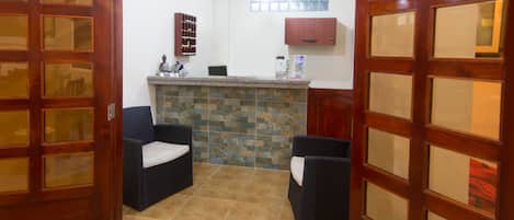 Habitación de hotel con piso de baldosas de piedra, puerta de madera con paneles de vidrio, un pequeño rincón de descanso con dos sillas, un mostrador de piedra y un armario de pared.