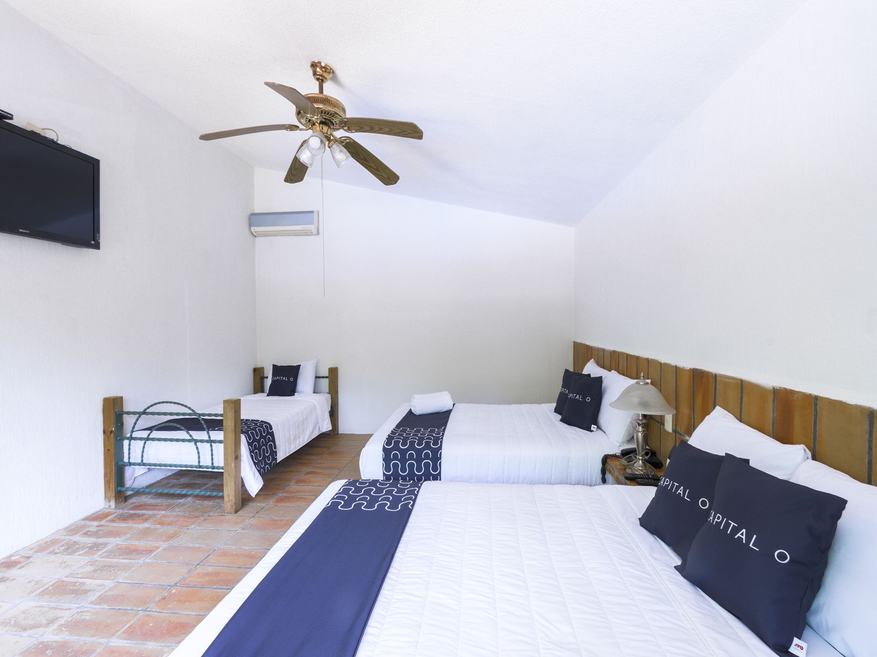 A hotel room with a large bed, two smaller beds, a ceiling fan, a TV, and a tiled floor.