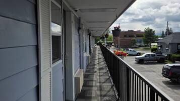 A covered outdoor corridor with a metal railing, a pathway, and a view of parked vehicles and buildings in the background.