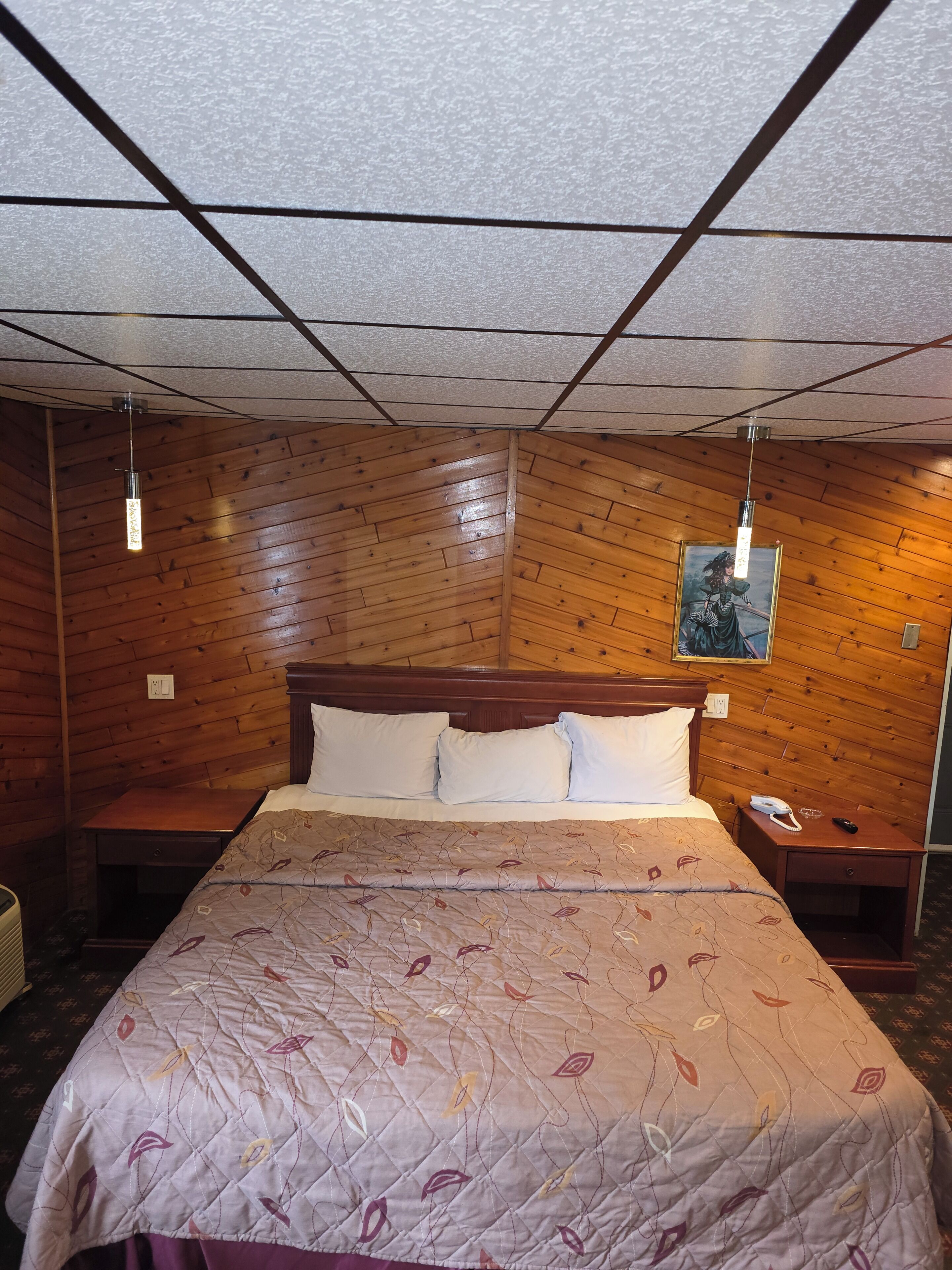 A hotel room with a wooden ceiling, a bed with a floral bedspread, a nightstand with a phone, and a painting on the wall.