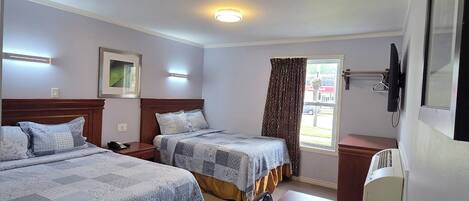 A hotel room with two beds, a wooden table, a desk, a chair, a window with curtains, a wall-mounted TV, and a ceiling light.