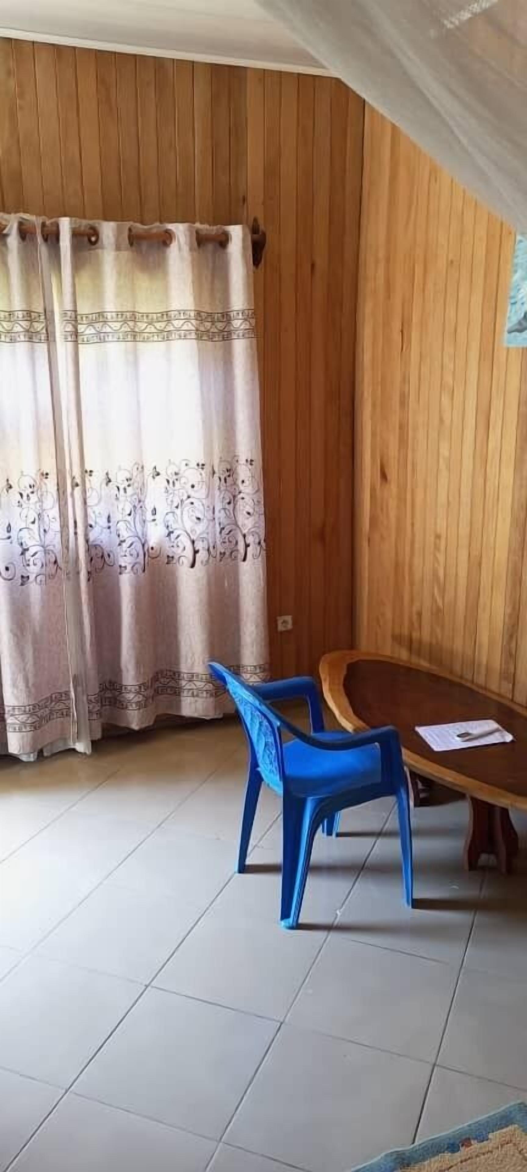 A room with wooden panel walls, a wooden table, two blue plastic chairs, and a patterned curtain.