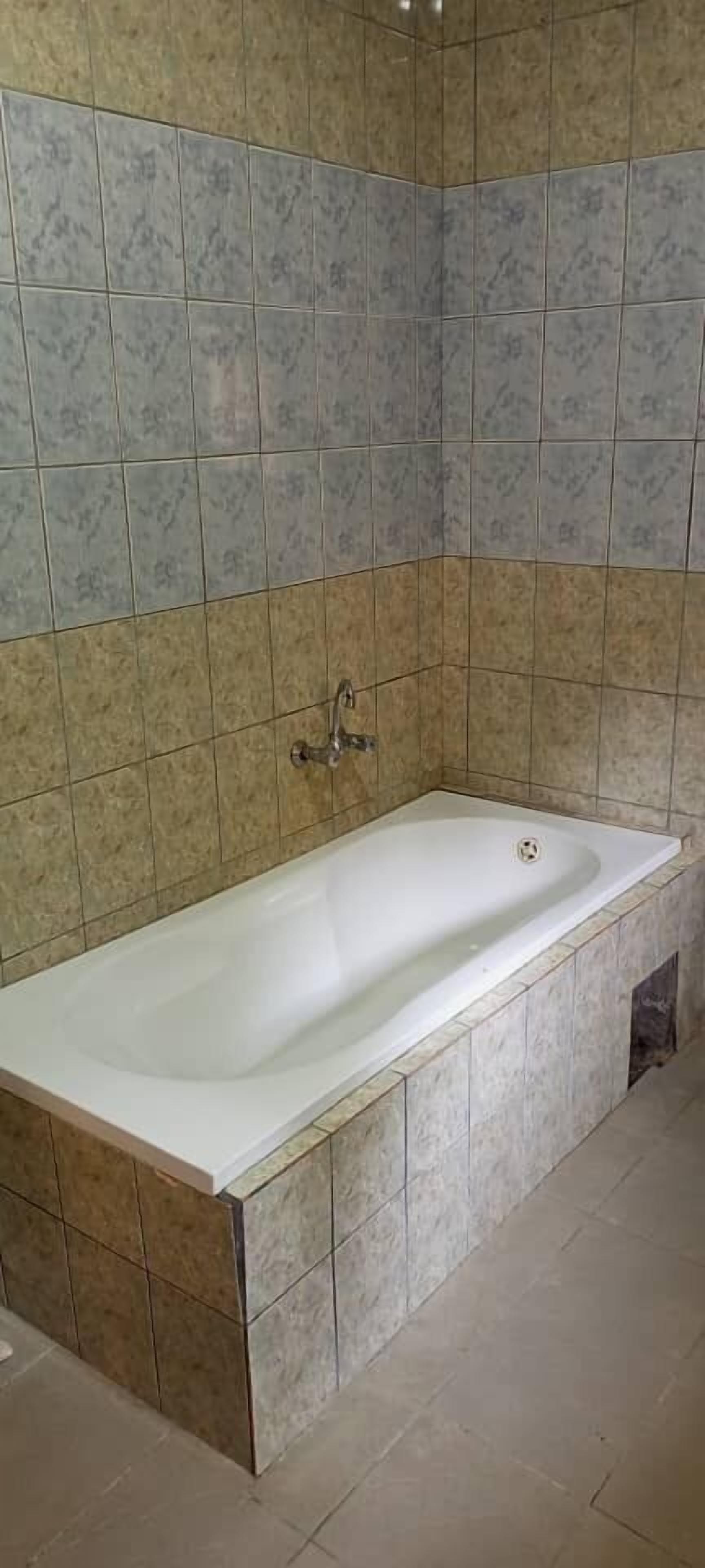 Eine standardmäßige, rechteckige Badewanne mit Chromarmatur vor einer gefliesten Wand mit einer Mischung aus beige- und grauen Fliesen.