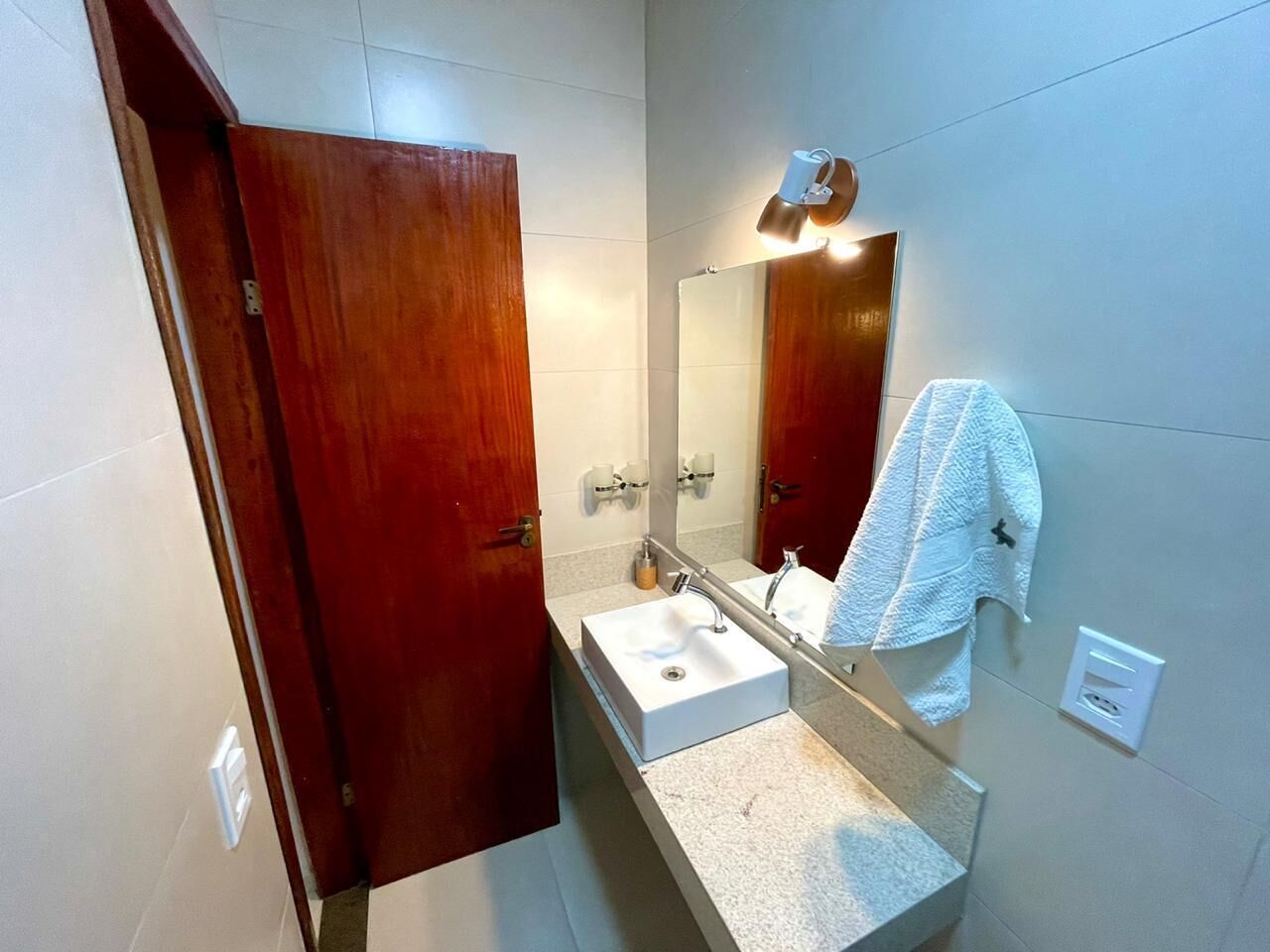 Ein Badezimmer mit Holztür, Waschbecken in Weiß, einem Spiegel und einem Handtuch, das an der Wand hängt.