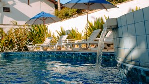 A swimming pool with water cascading into it, surrounded by lounge chairs and umbrellas, with a building in the background. - HOTELARE Hotel Brunello (Paraty)