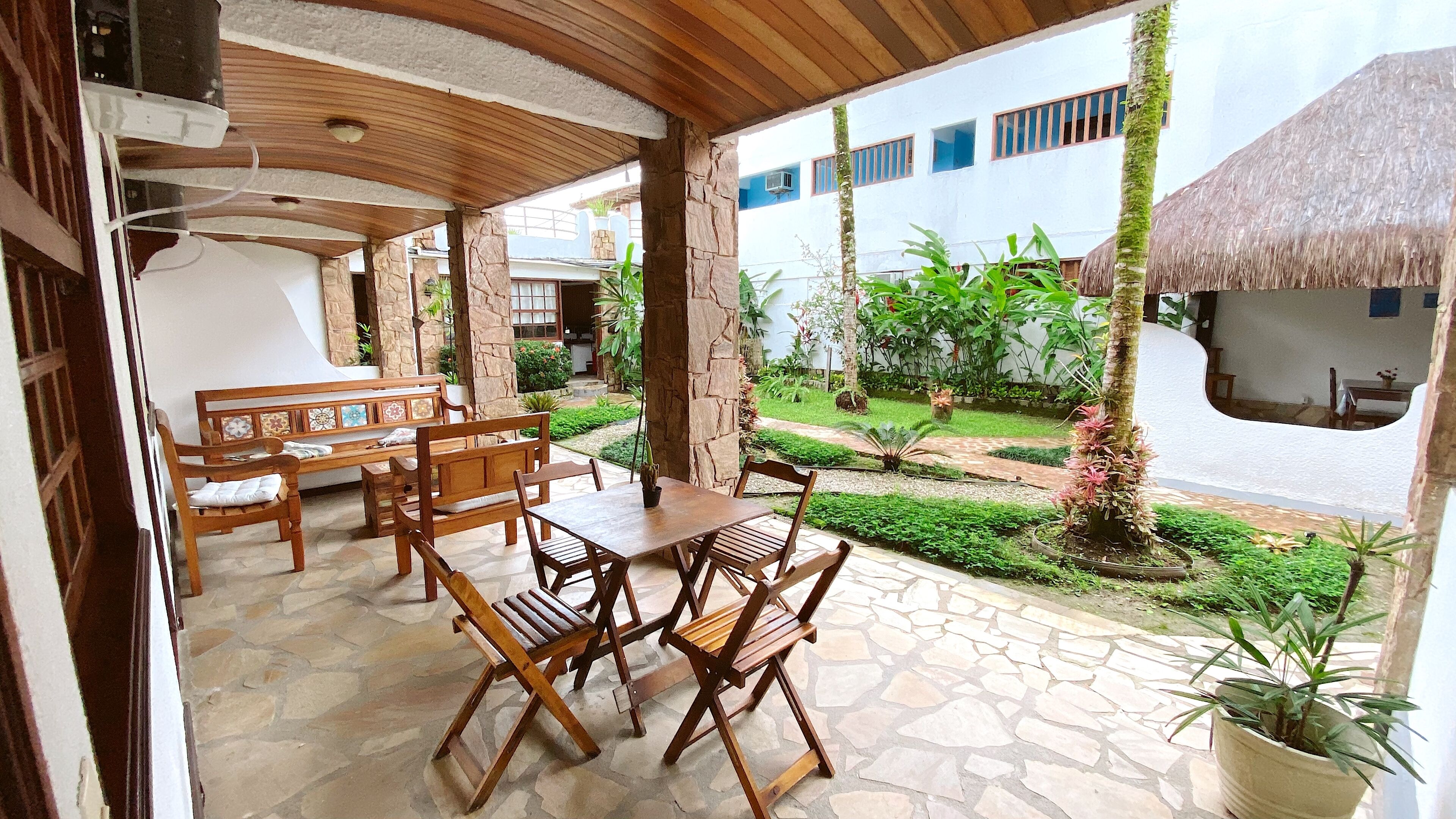 An outdoor patio with wooden furniture and a stone floor, surrounded by greenery and a view of a building with a blue and white facade.