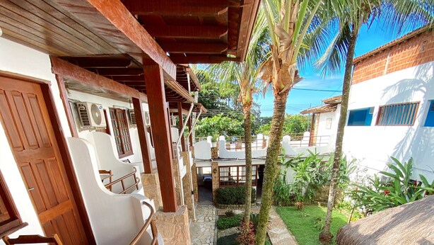 A covered outdoor seating area with wooden furniture and a stone pathway leading to a garden with palm trees and a thatched roof structure. - HOTELARE Hotel Brunello (Paraty)
