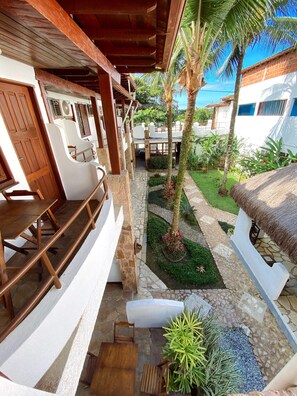 A covered outdoor seating area with wooden furniture and a stone pathway leading to a garden with palm trees and a thatched roof structure. - HOTELARE Hotel Brunello (Paraty)
