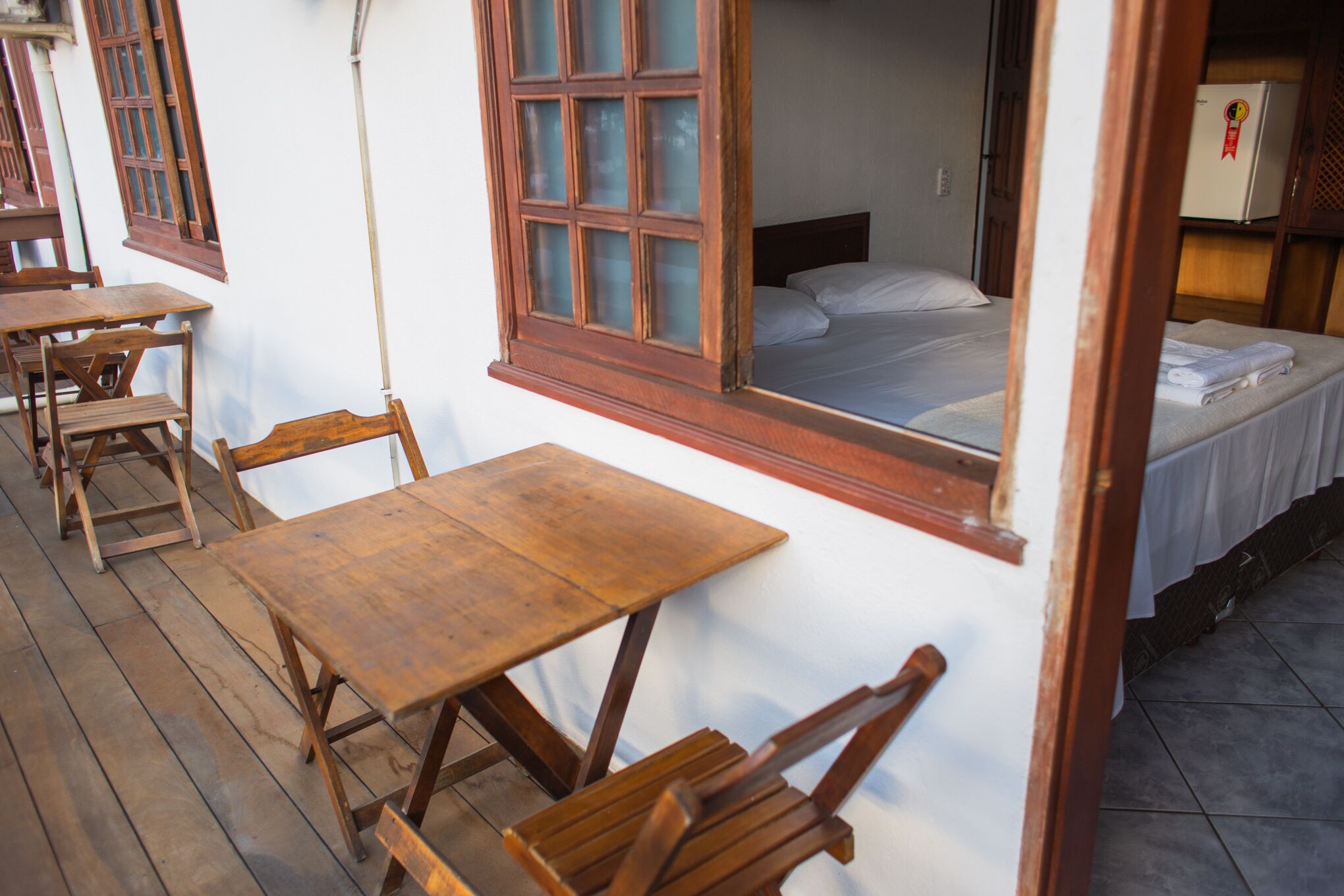 A room with a wooden table and chairs, a bed with white linens, and a window with wooden frames.