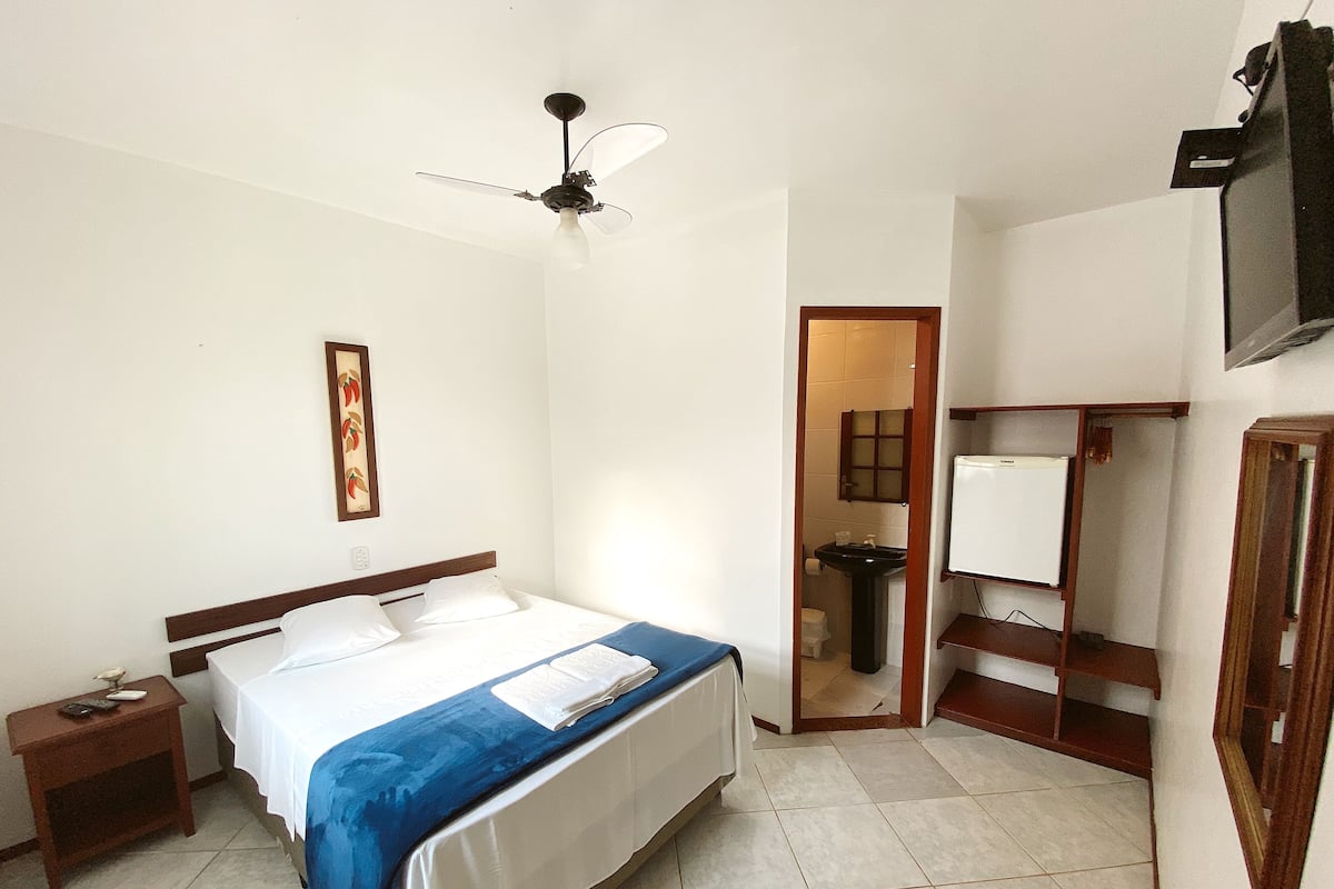 Habitación de hotel con cama, mesita de noche, ventilador de techo, baño visible a través de una puerta abierta y televisor montado en la pared.