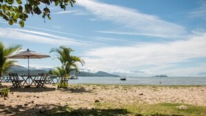A beach with a table and chairs under a canopy, palm trees, and a view of the sea and mountains. - HOTELARE Hotel Brunello (Paraty)