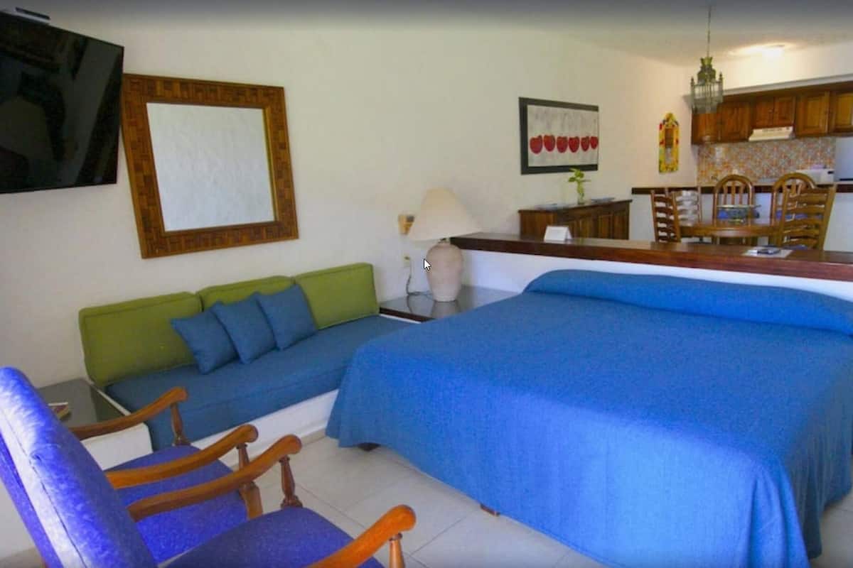 Una habitación con una cama, un sofá verde, una silla de madera, una mesita y una zona de cocina con armarios de madera y un lavamanos.