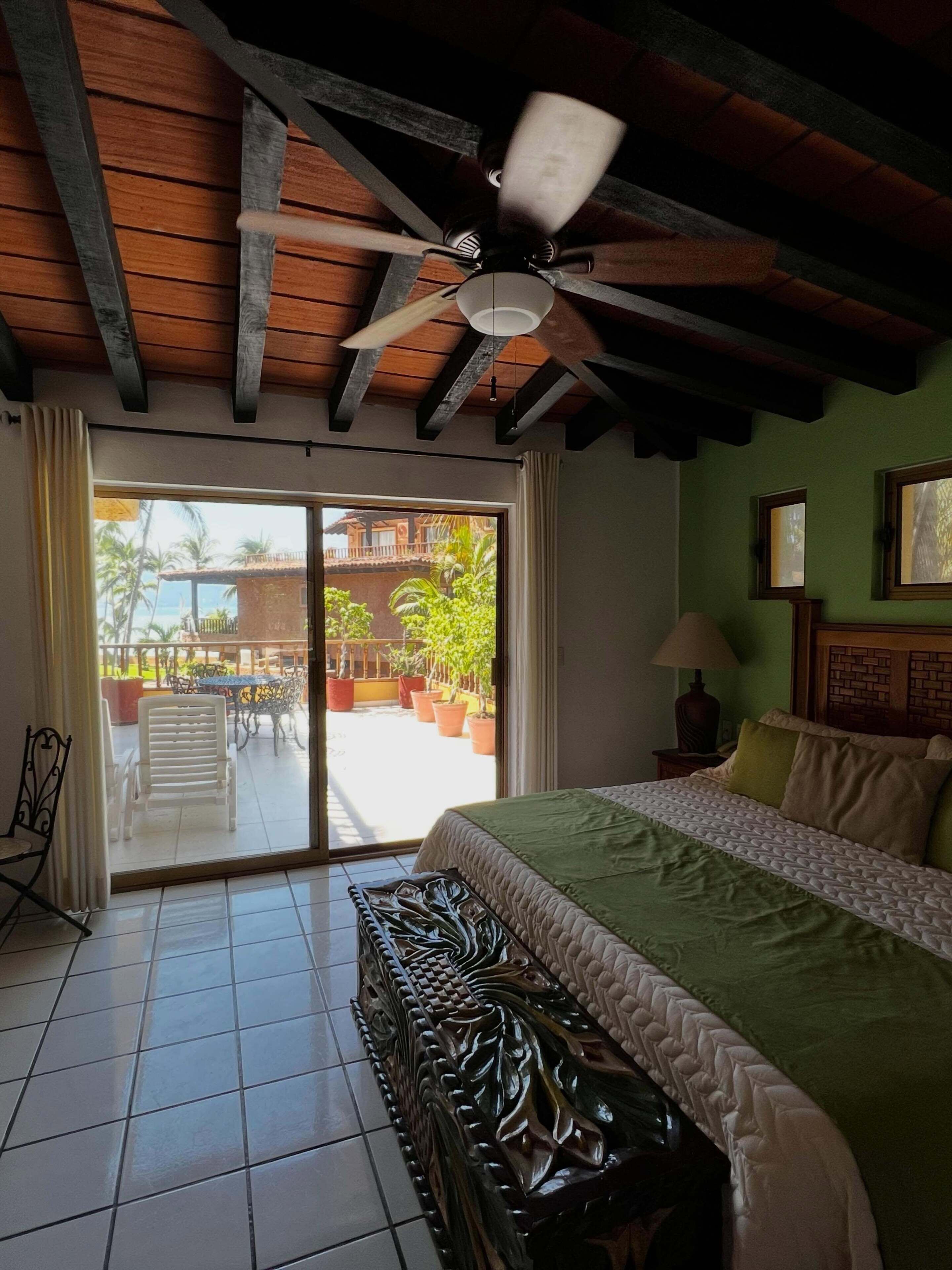 Camera d'albergo con un letto, una ventola a soffitto, vista all'esterno e un balcone con sedie e piante in vaso.