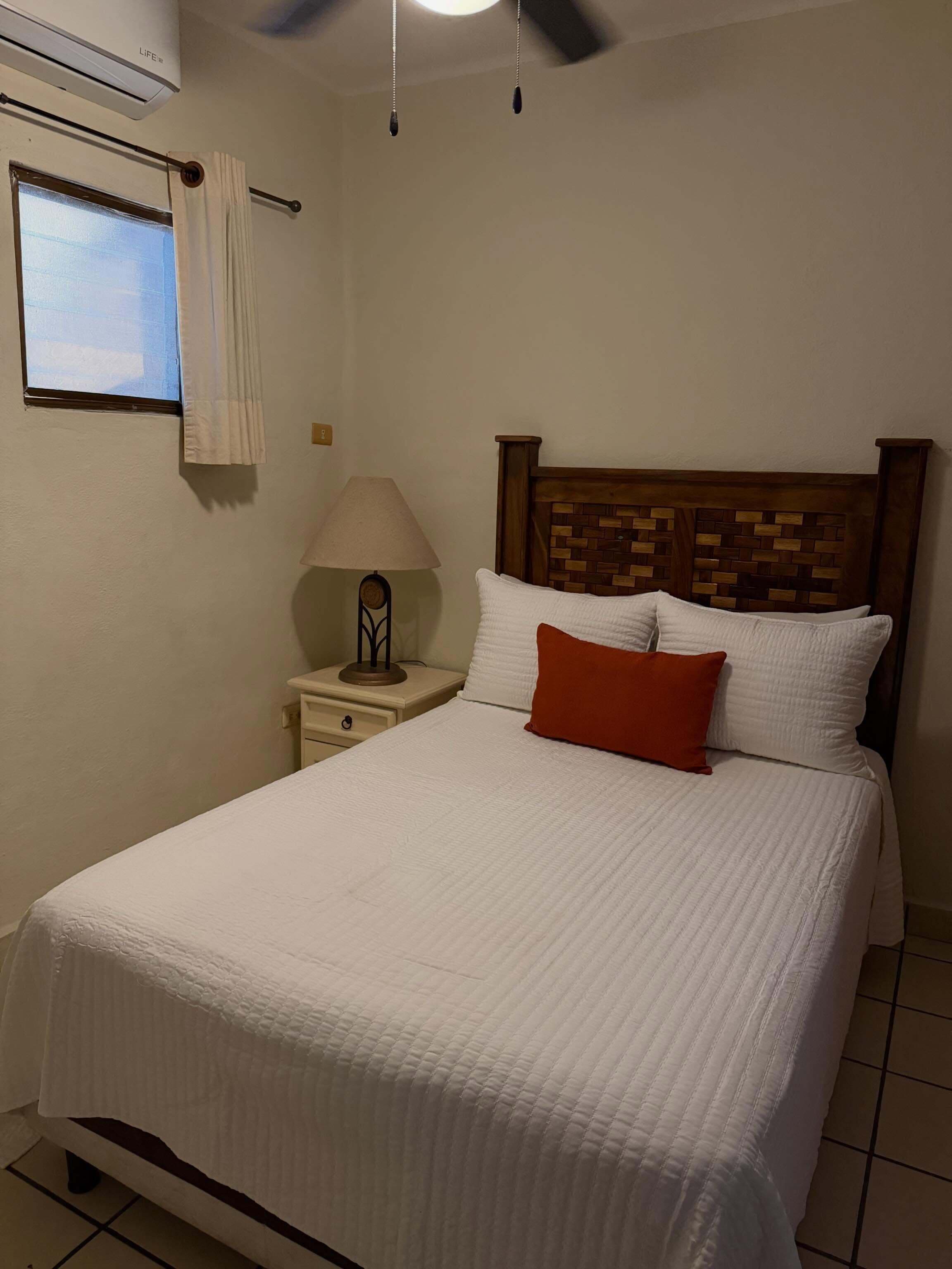 A bedroom with a bed, a nightstand, a lamp, a window with curtains, and an air conditioning unit.