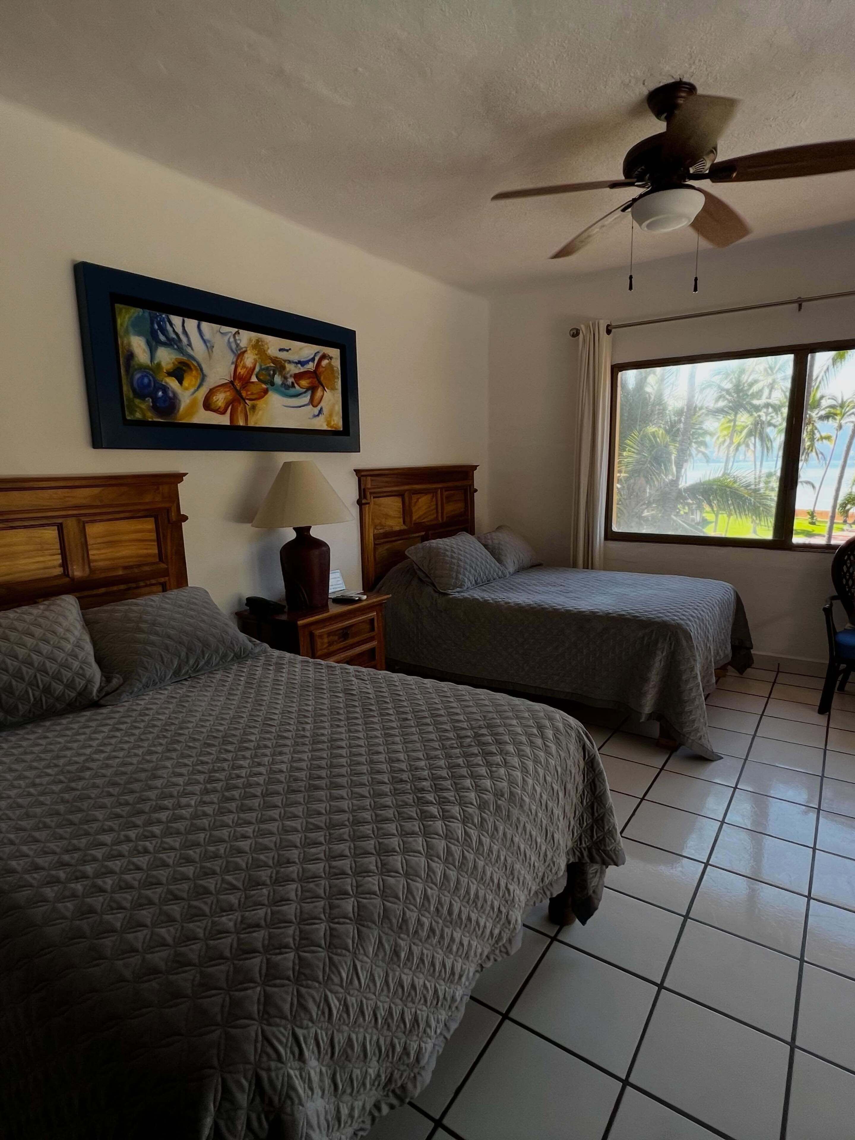 A hotel room with two beds, a ceiling fan, a window with a view of palm trees, and a painting on the wall.