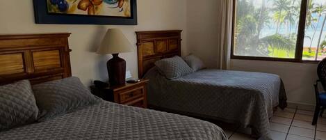 A hotel room with two beds, a ceiling fan, a window with a view of palm trees, and a painting on the wall.