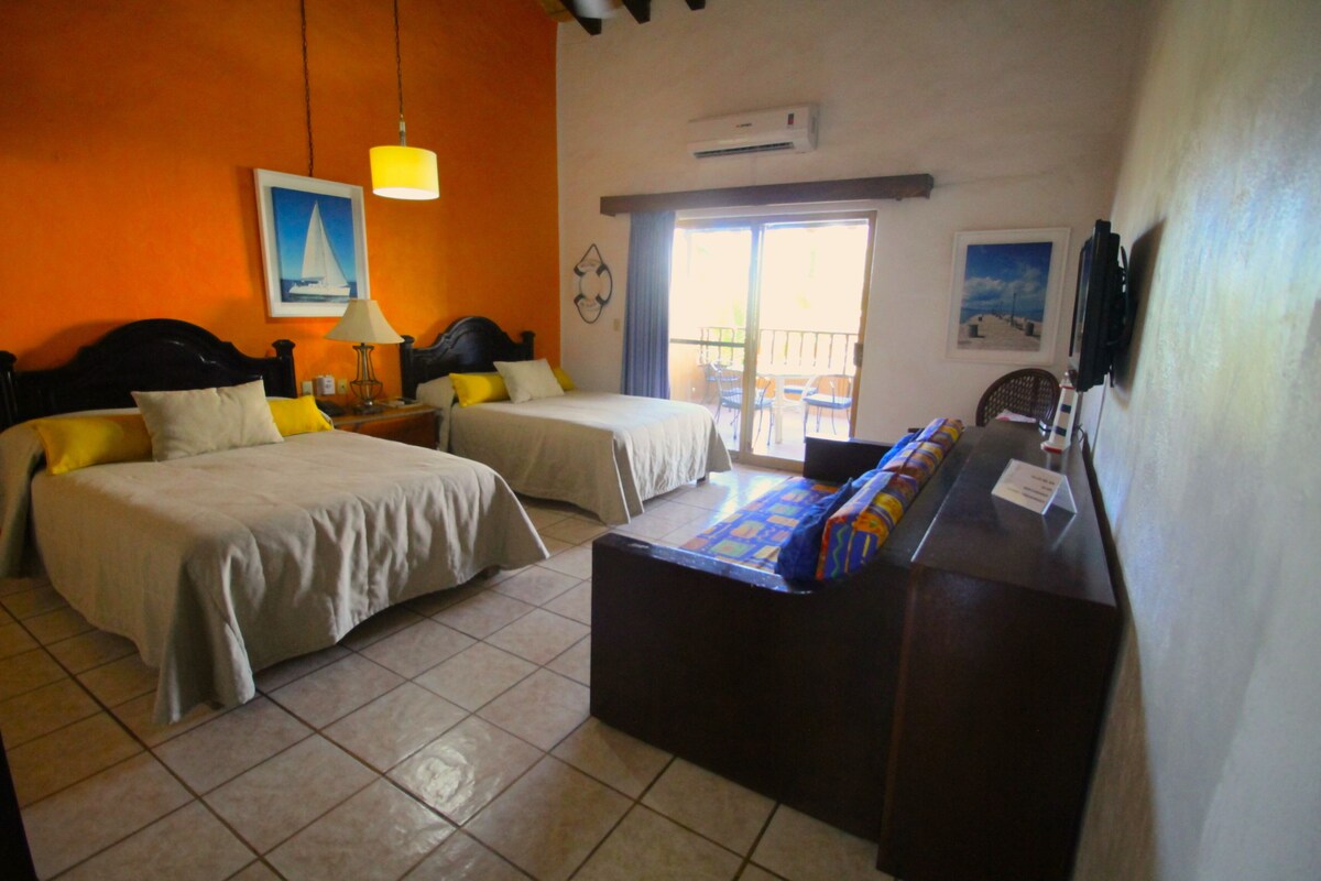 A hotel room with two beds, a sofa, a TV, and a balcony with chairs.