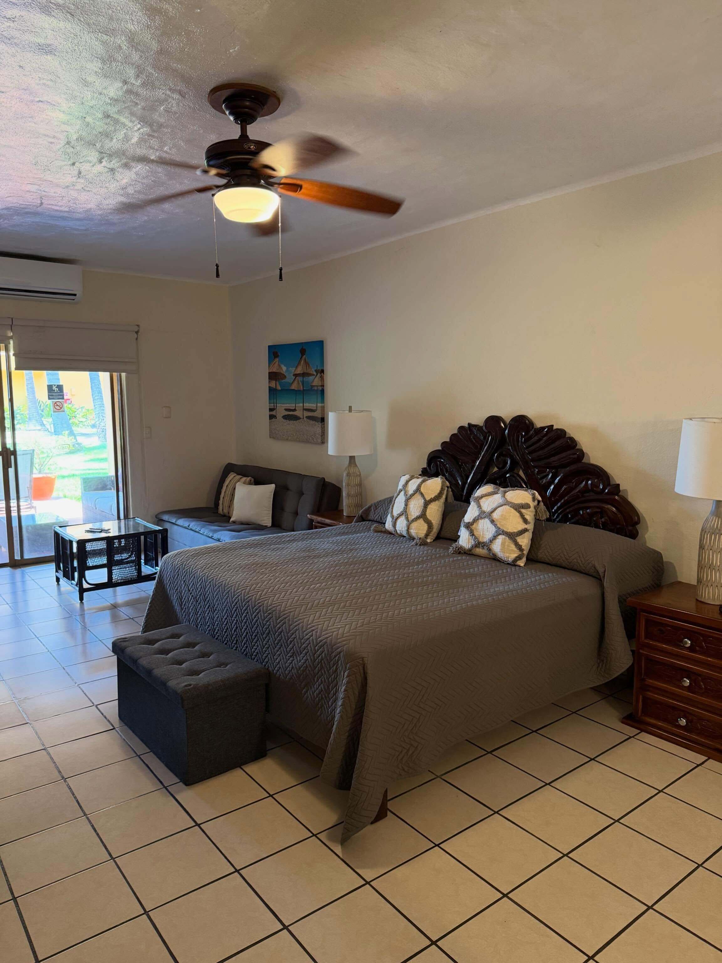 A hotel room with a bed, a desk, a chair, a ceiling fan, and a painting on the wall.