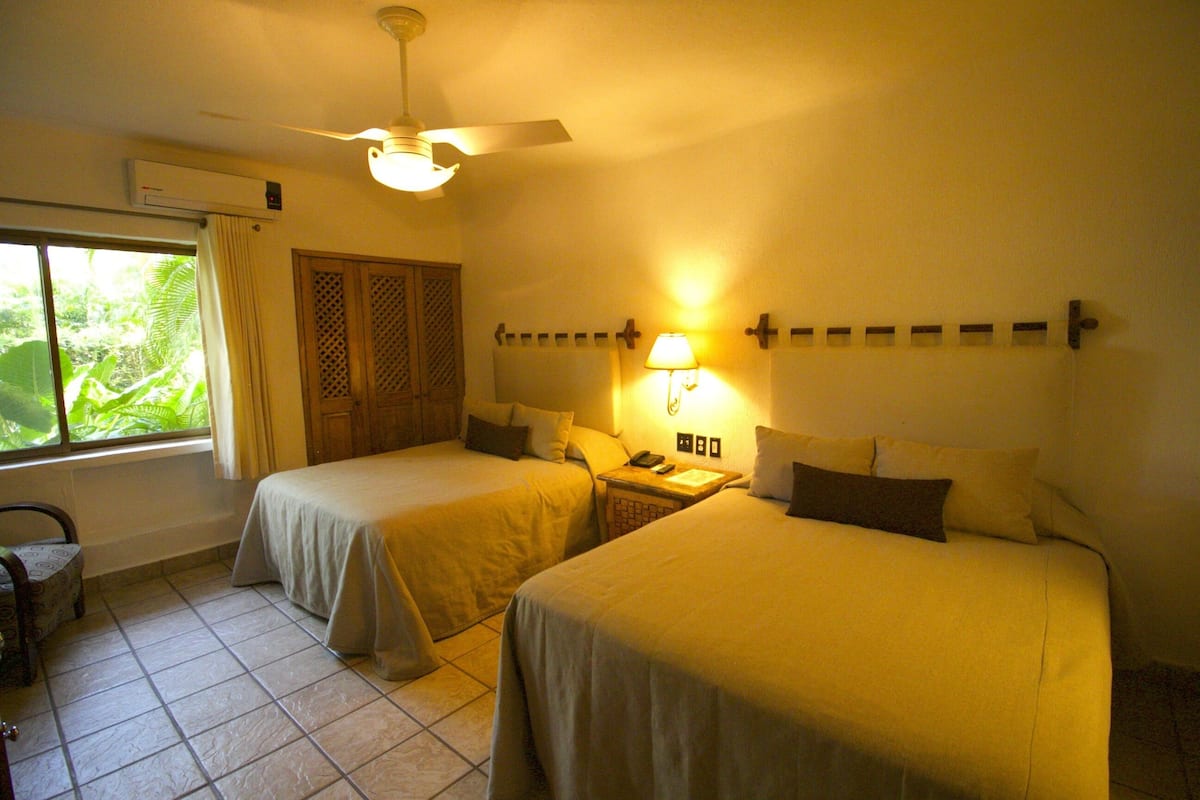Una habitación de hotel con dos camas, un ventilador de techo, una mesita de noche con lámpara y una ventana con cortinas.