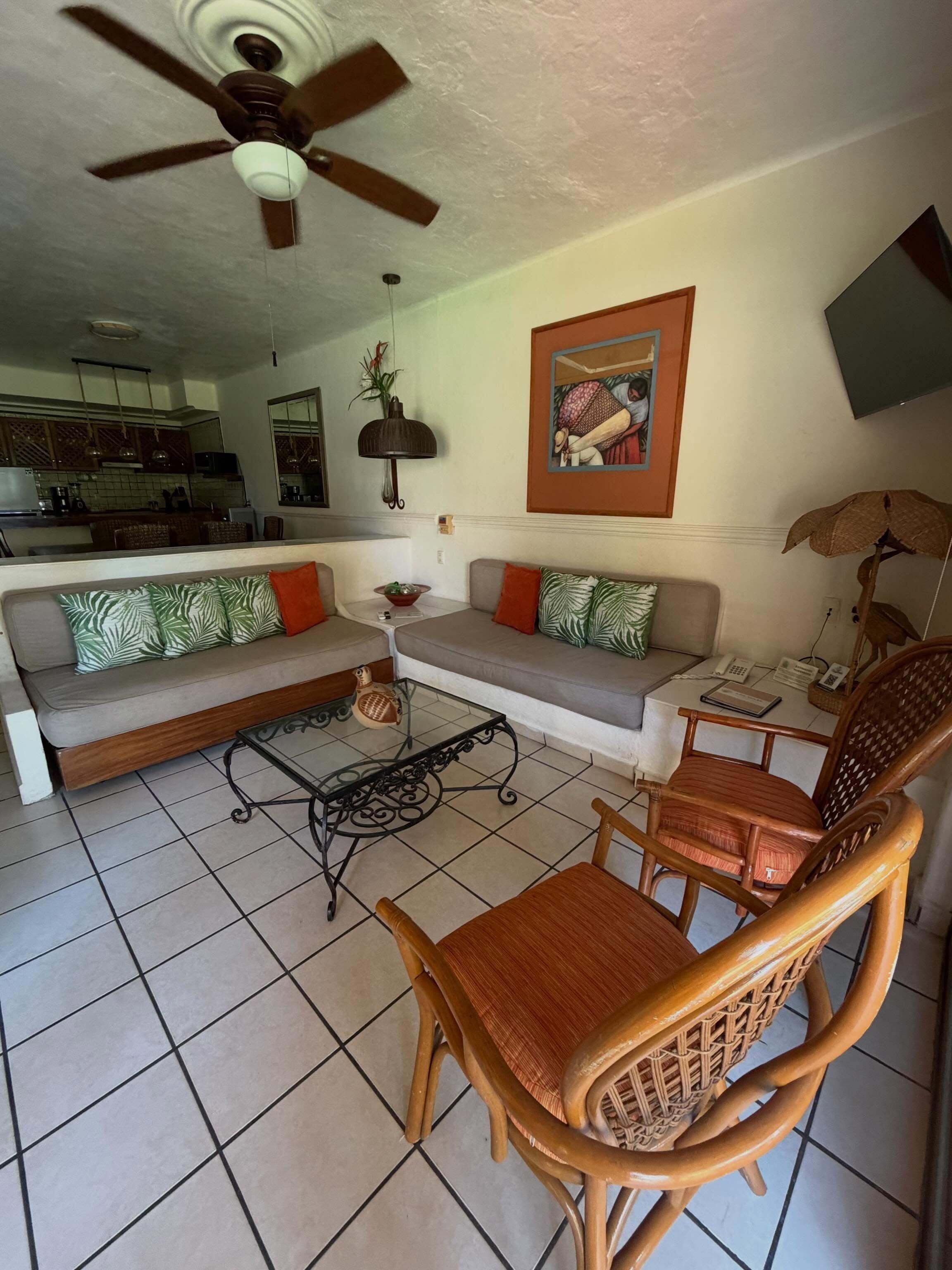 A living room with a sofa, two armchairs, a coffee table, a ceiling fan, a kitchen in the background, and a framed picture on the wall.
