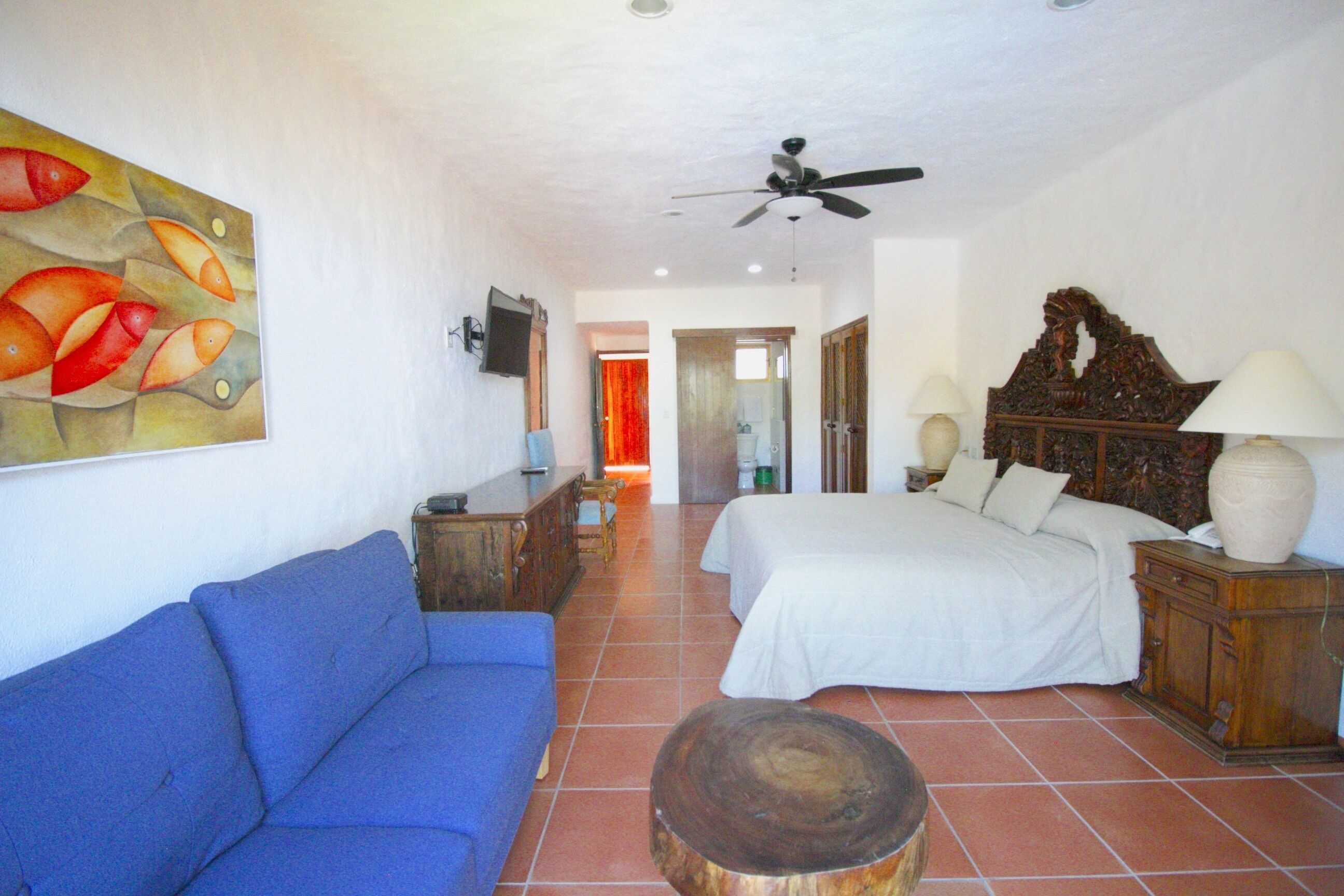 A hotel room with a blue sofa, two beds, a ceiling fan, a painting, and a wooden cabinet.