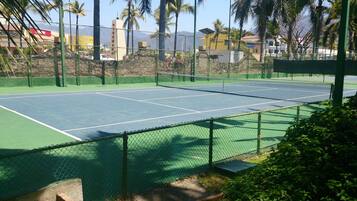 Una cancha de tenis al aire libre con superficie verde, rodeada por una cerca de malla metálica.