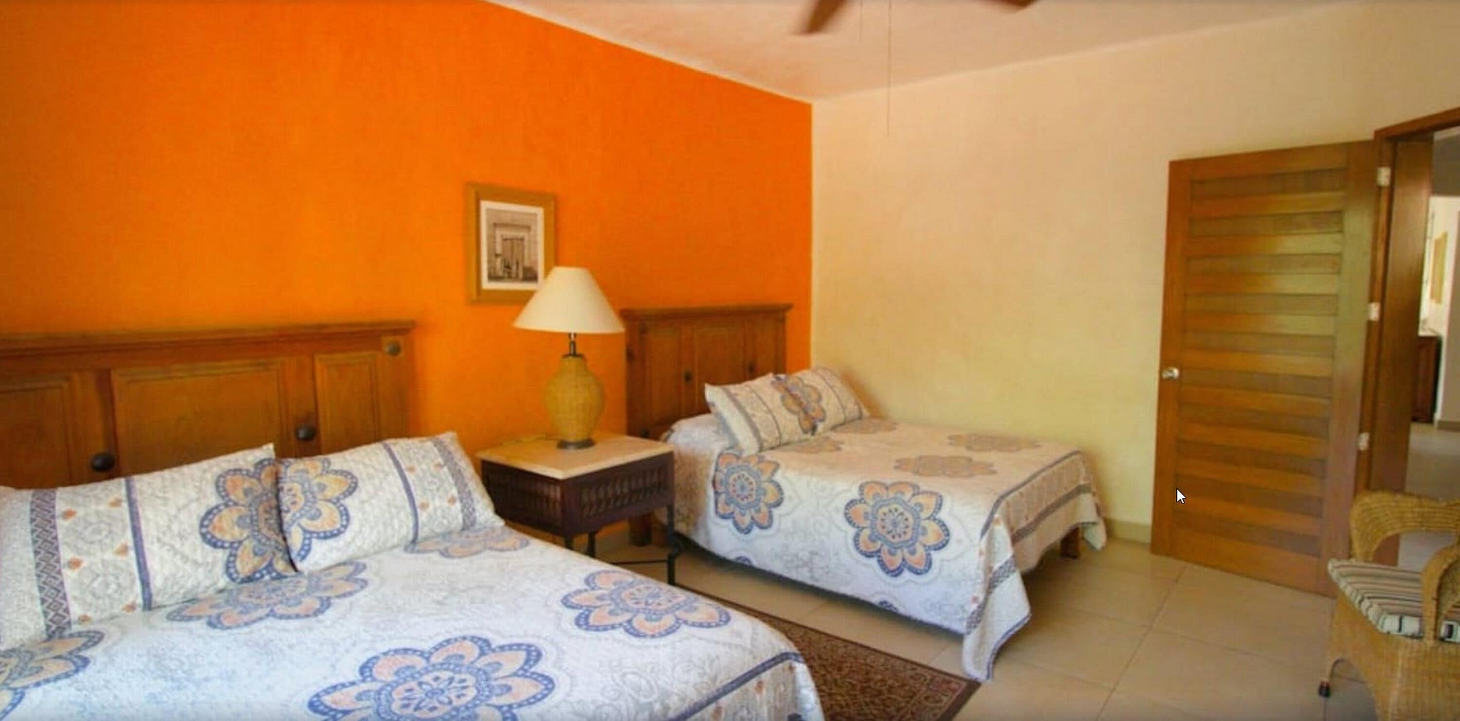 A hotel room with two beds, a wooden headboard, a nightstand with a lamp, and an orange wall.