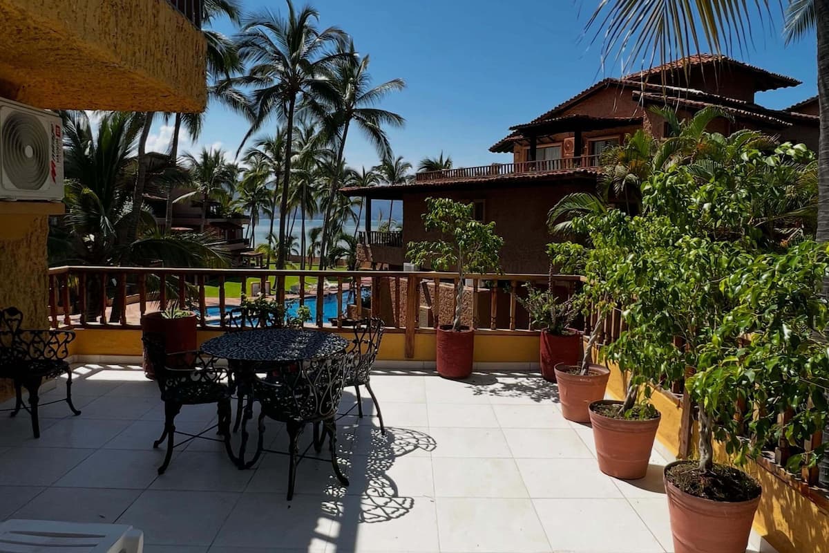 A patio area with a pool, outdoor furniture, potted plants, and a building with a balcony.
