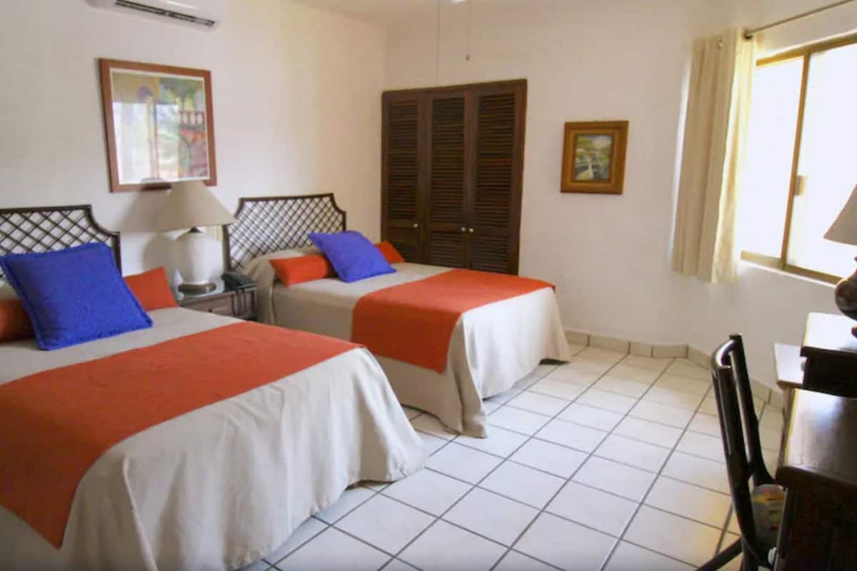 Habitación de hotel con dos camas, cada una con cubrecama rojo y blanco, una mesita de noche con lámpara, un armario de madera y una ventana con cortinas.