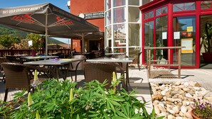 An outdoor seating area with wicker chairs and tables, a red building with glass windows, and a sign for a hotel-restaurant. - Le Clos du Hahnenberg (Urmatt)