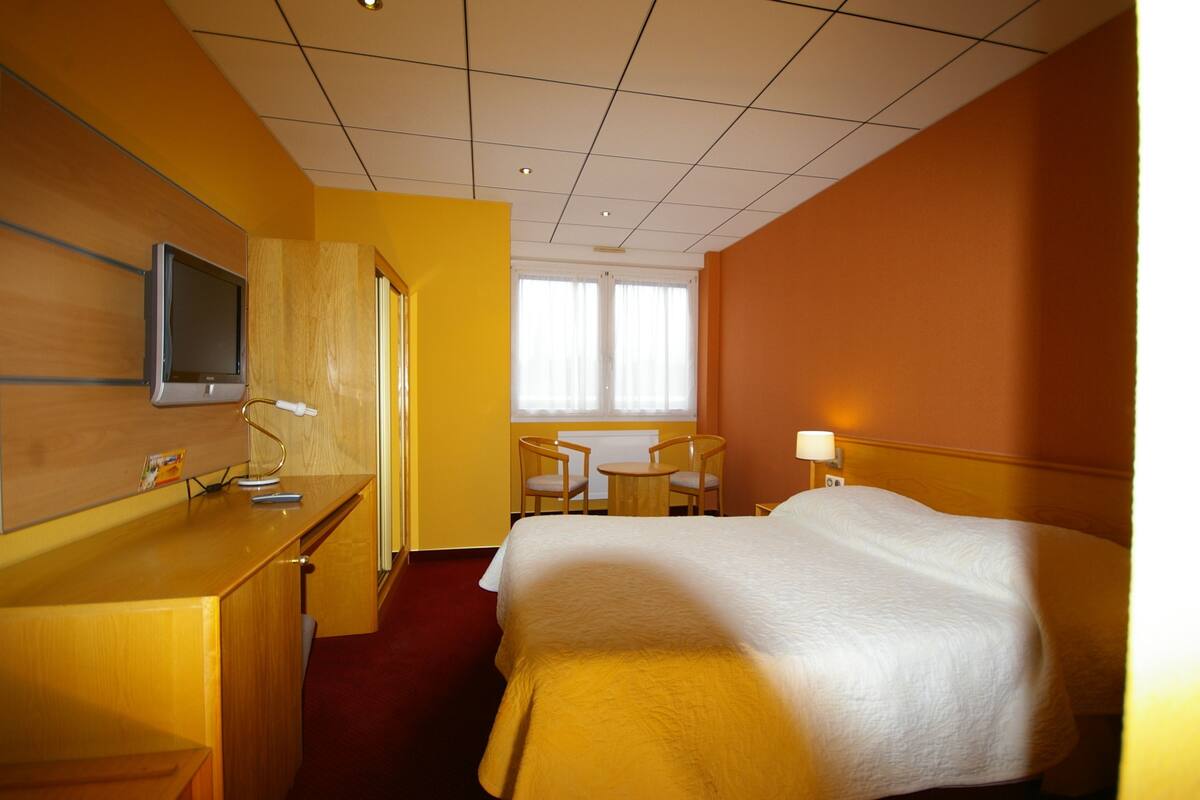 A hotel room with a bed, a desk with a television, a small table and chairs, and a window with curtains.