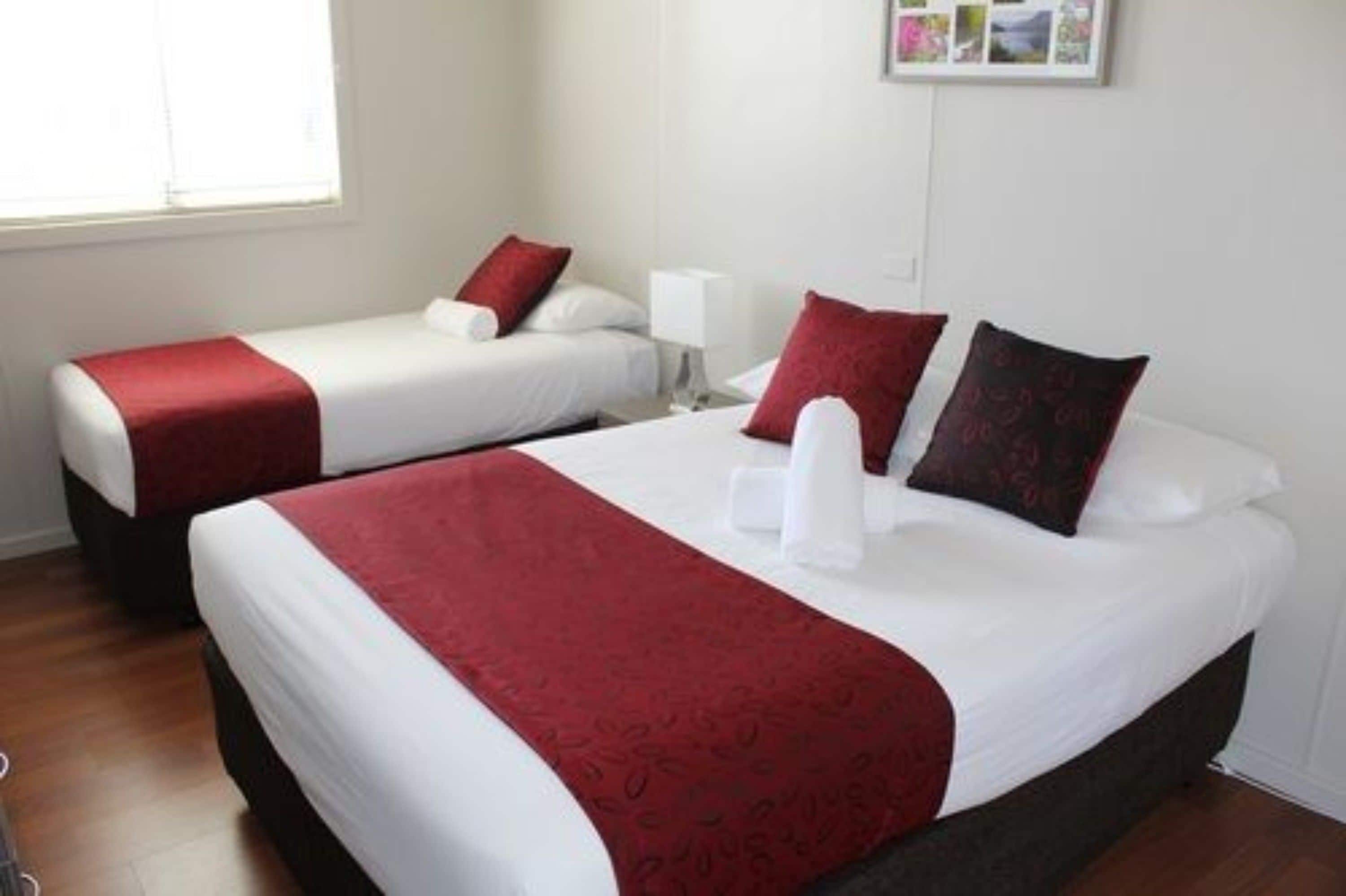 Une chambre d’hôtel avec deux lits, chacun recouvert d’une couverture rouge et doté de draps blancs. On y trouve une petite table de chevet avec une lampe, et un cadre avec une photo accroché au mur.