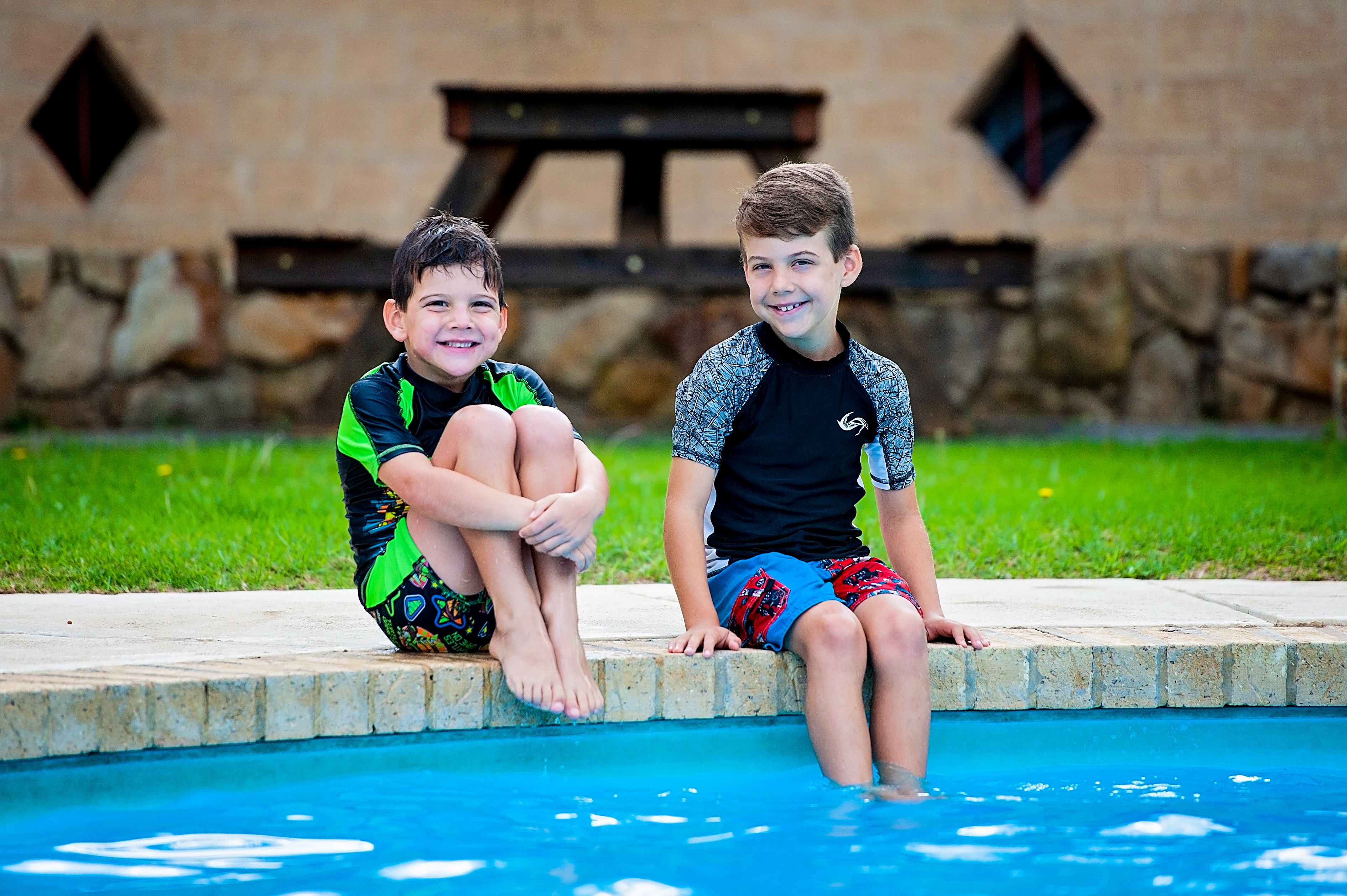 Two children sitting by a pool with a stone wall and a picnic table in the background.