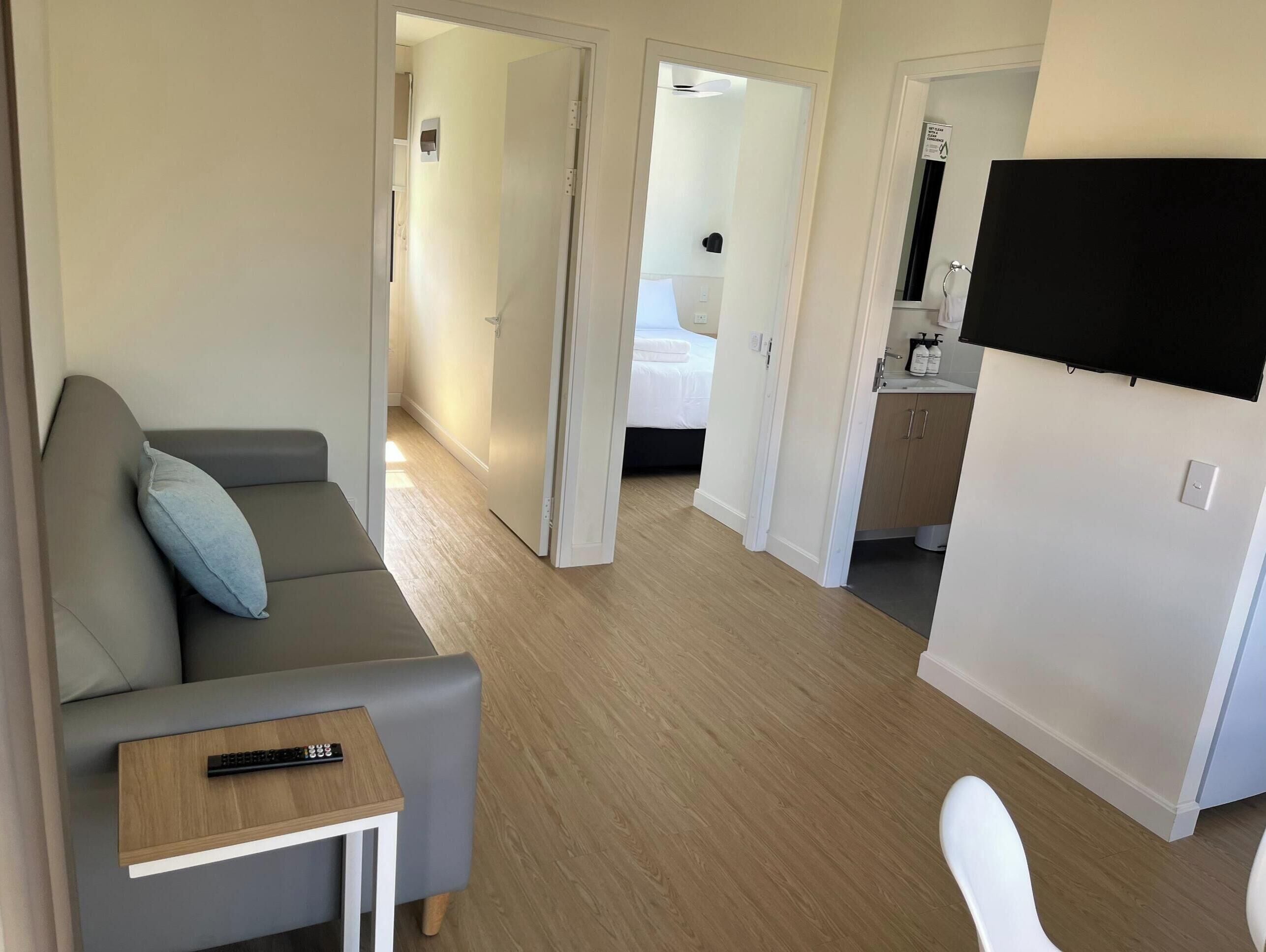 A modern hotel room with a grey sofa, a small table with a remote control, a TV, and a bathroom visible through an open door.