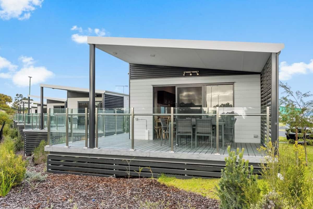 Une maison moderne de plain-pied, dotée d’une terrasse et d’une balustrade en verre, et entourée de verdure.