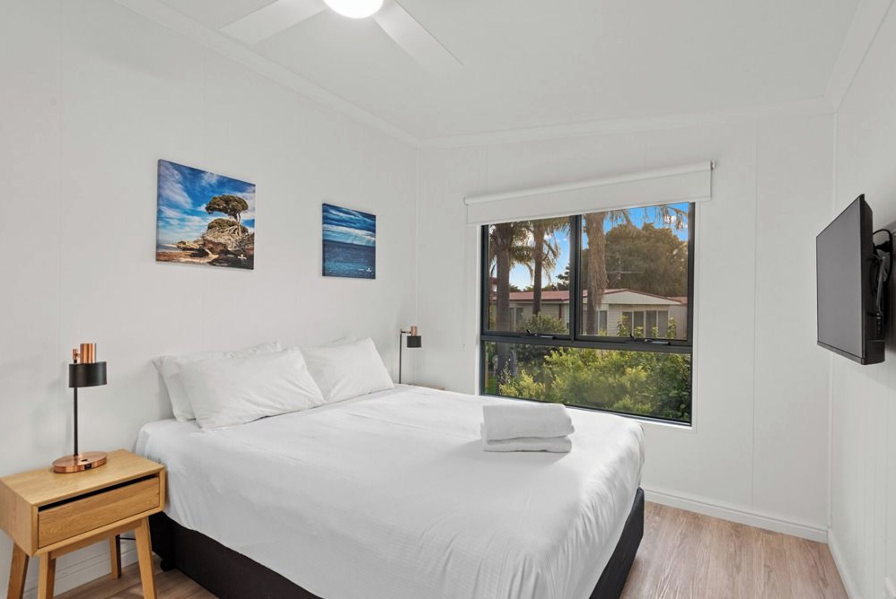 A bedroom with a bed, a nightstand, a lamp, two paintings, a window with a view, and a TV mounted on the wall.
