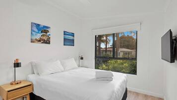 A bedroom with a bed, a nightstand, a lamp, two paintings, a window with a view, and a TV mounted on the wall.