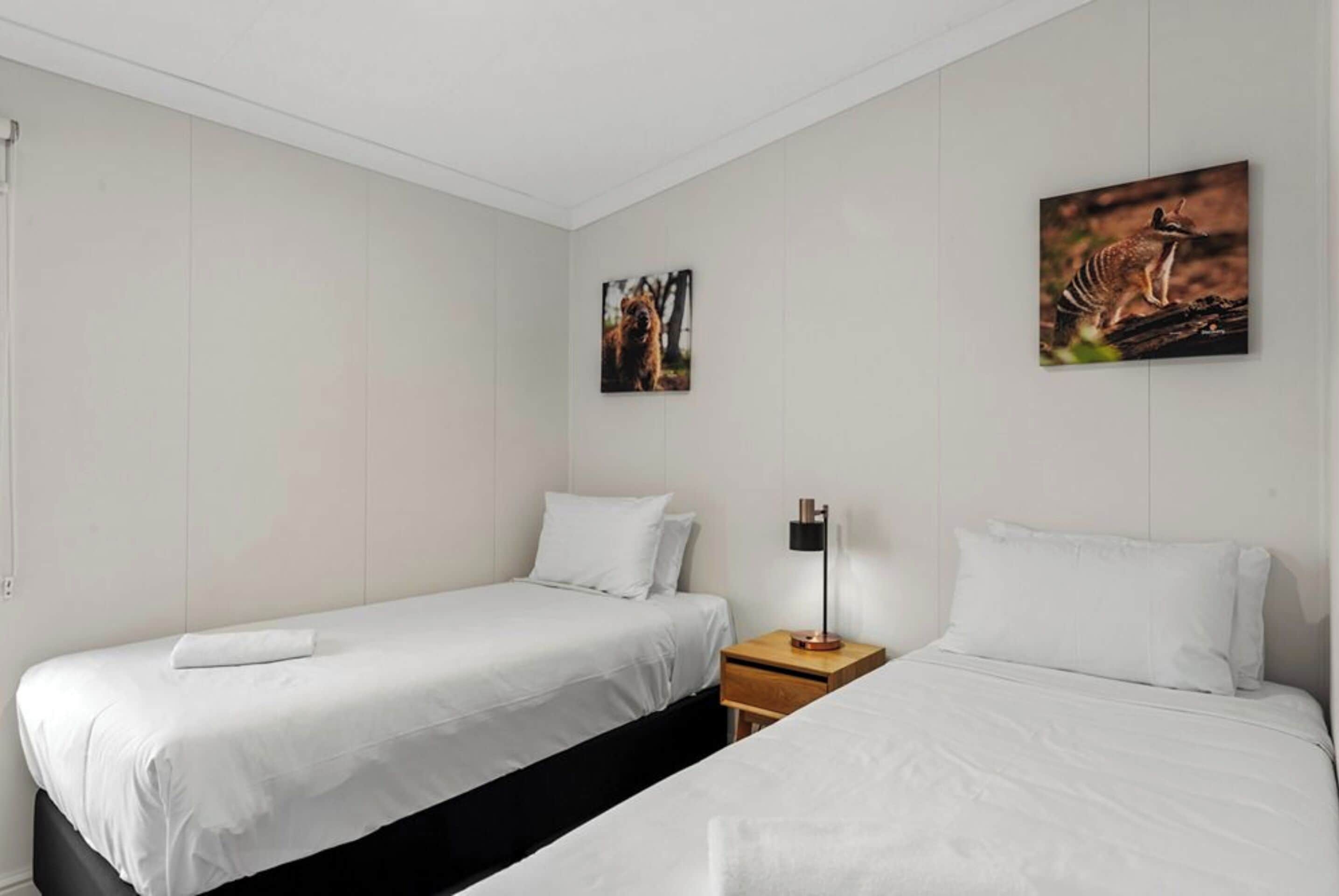 A hotel room with two beds, white bedding, a nightstand with a lamp, and two framed pictures on the wall.