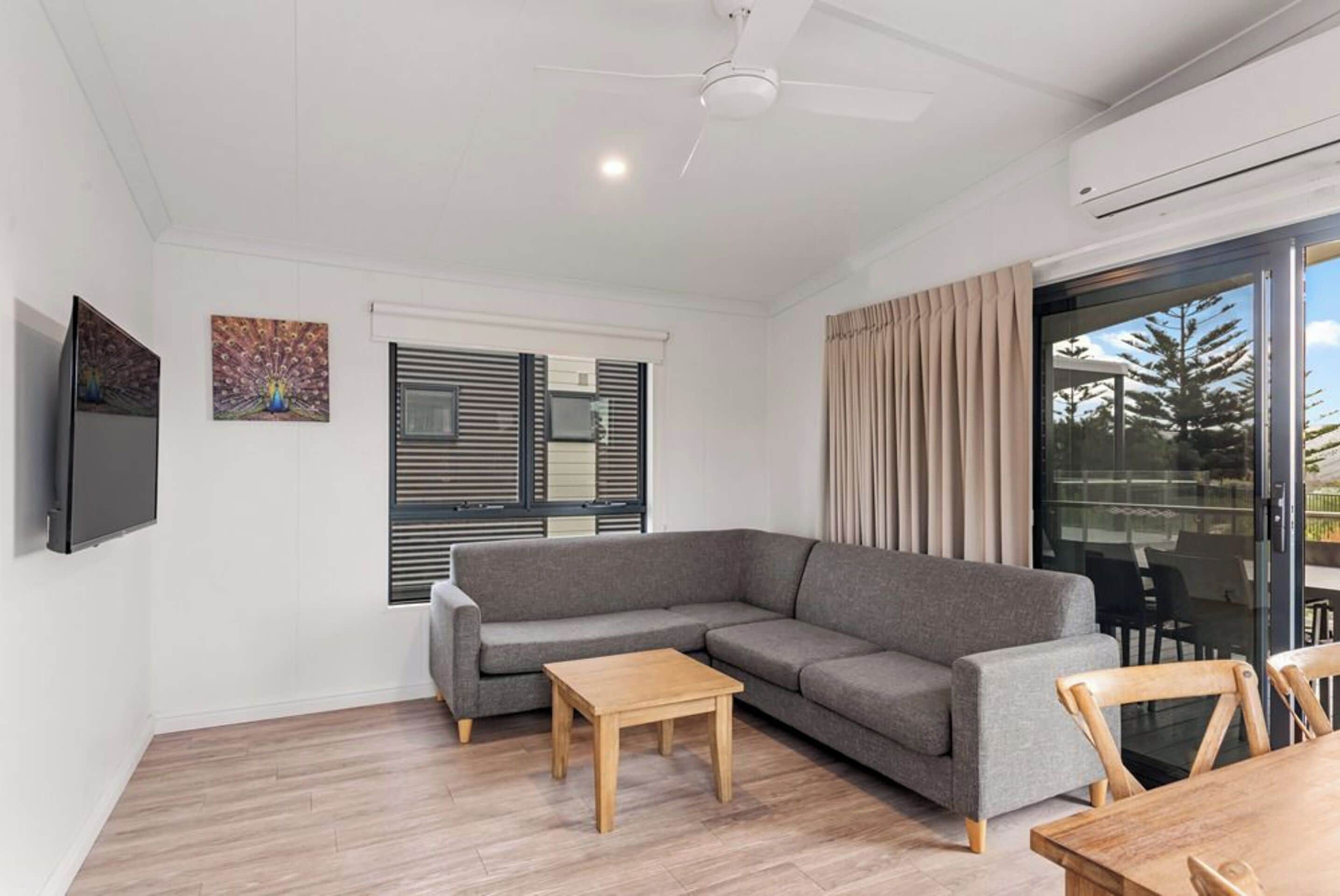A modern living room with a grey sectional sofa, a wooden coffee table, a flat-screen TV mounted on the wall, and a sliding glass door leading to an outdoor area.