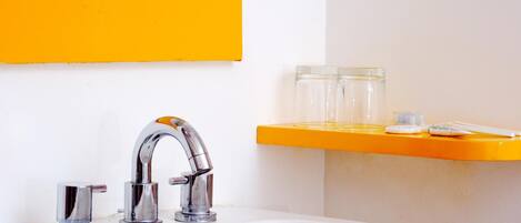 Un baño con un lavabo blanco, grifos cromados y un estante amarillo con recipientes de vidrio y un cepillo de dientes.