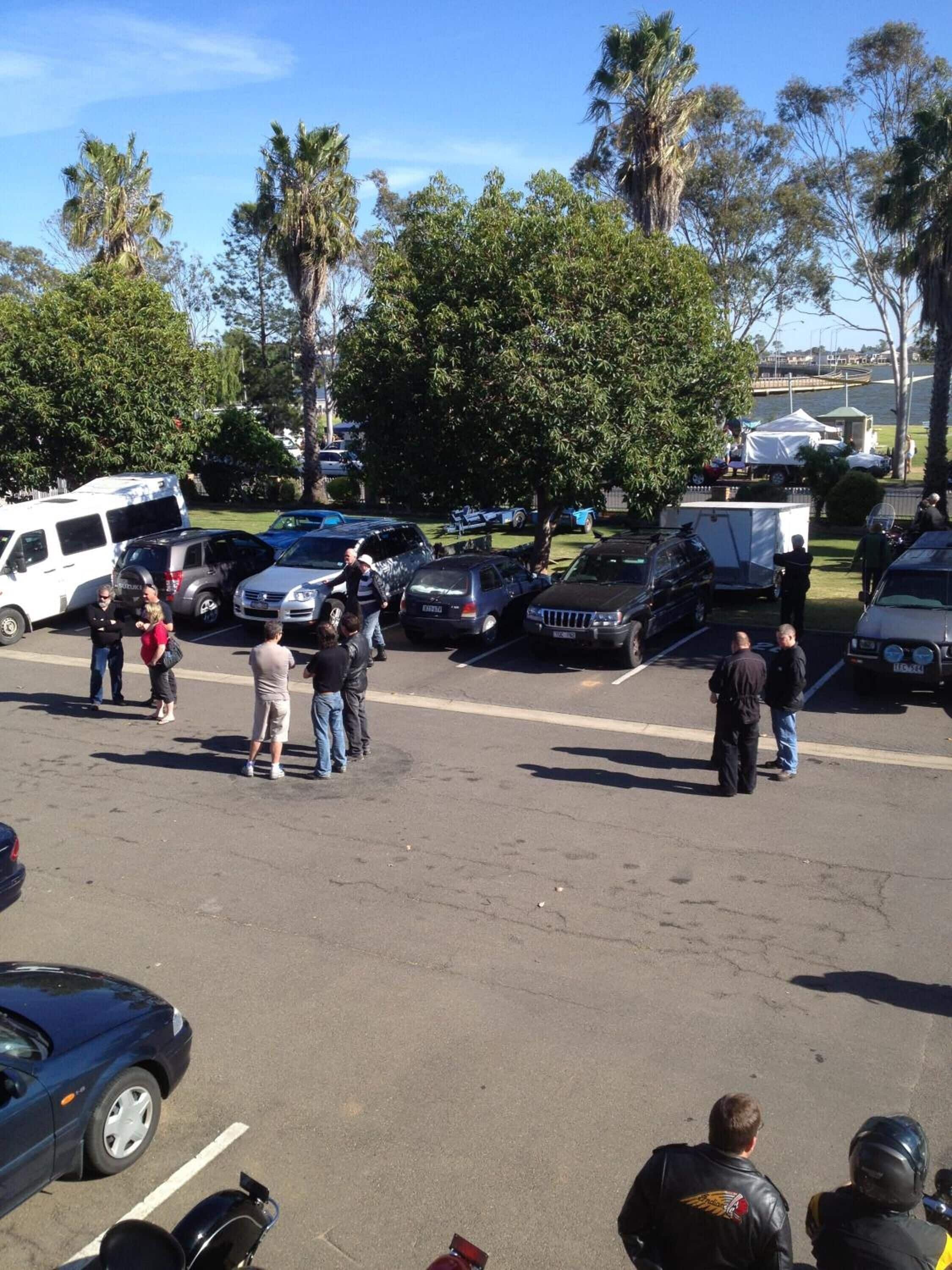 A parking lot with several vehicles, including cars and a van, and a group of people standing and conversing.