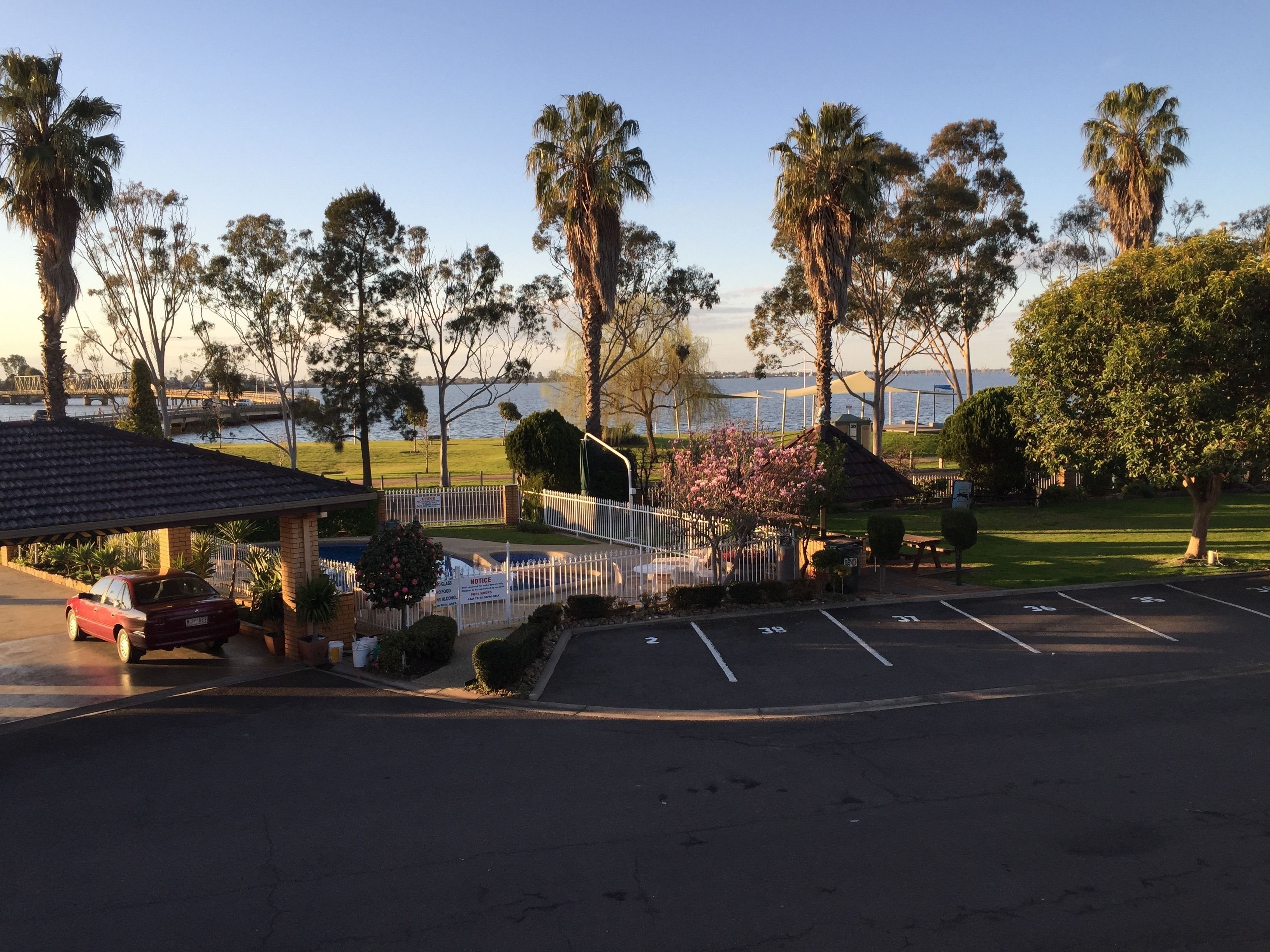 A parking lot with a covered area, a red car, and a view of a body of water with trees and a bridge in the background.